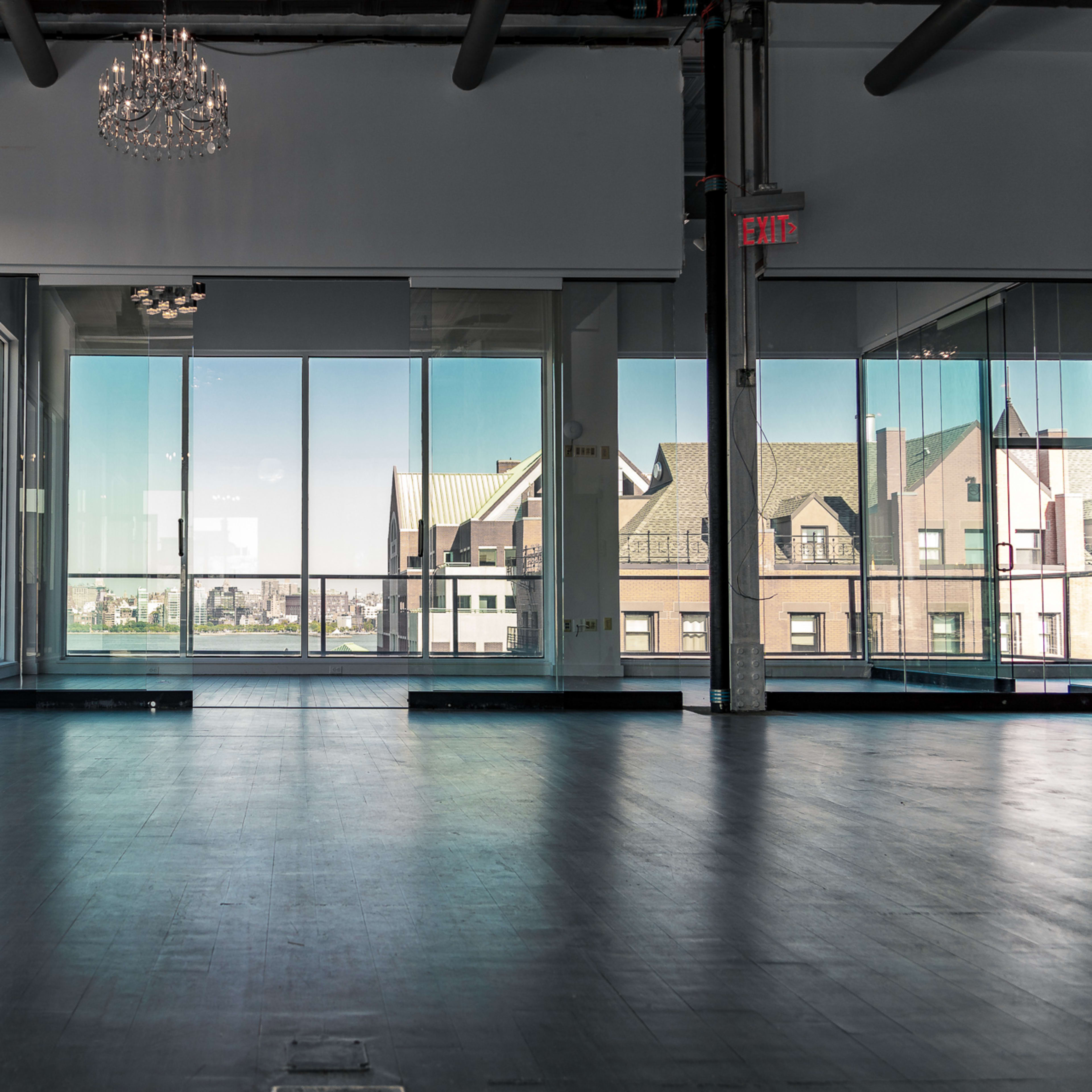 A spacious interior room features large windows overlooking city buildings and a chandelier above the empty wooden floor.