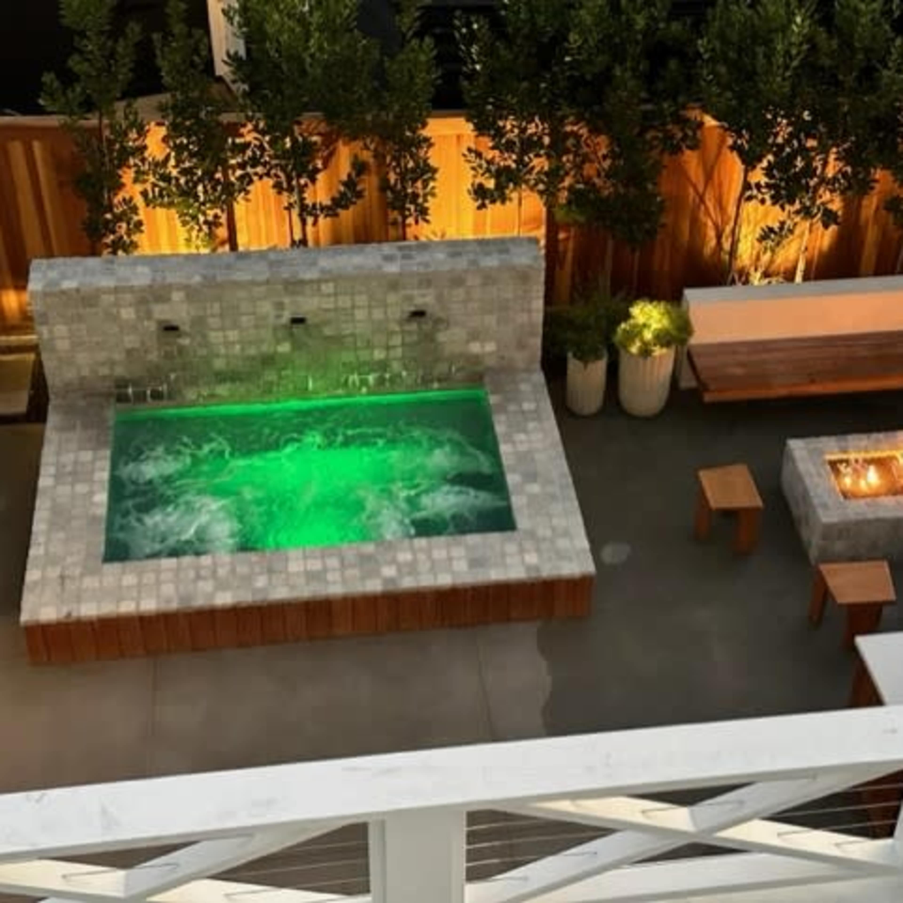 A modern outdoor patio featuring a hot tub with green lighting and a stone fire pit surrounded by wooden seating.