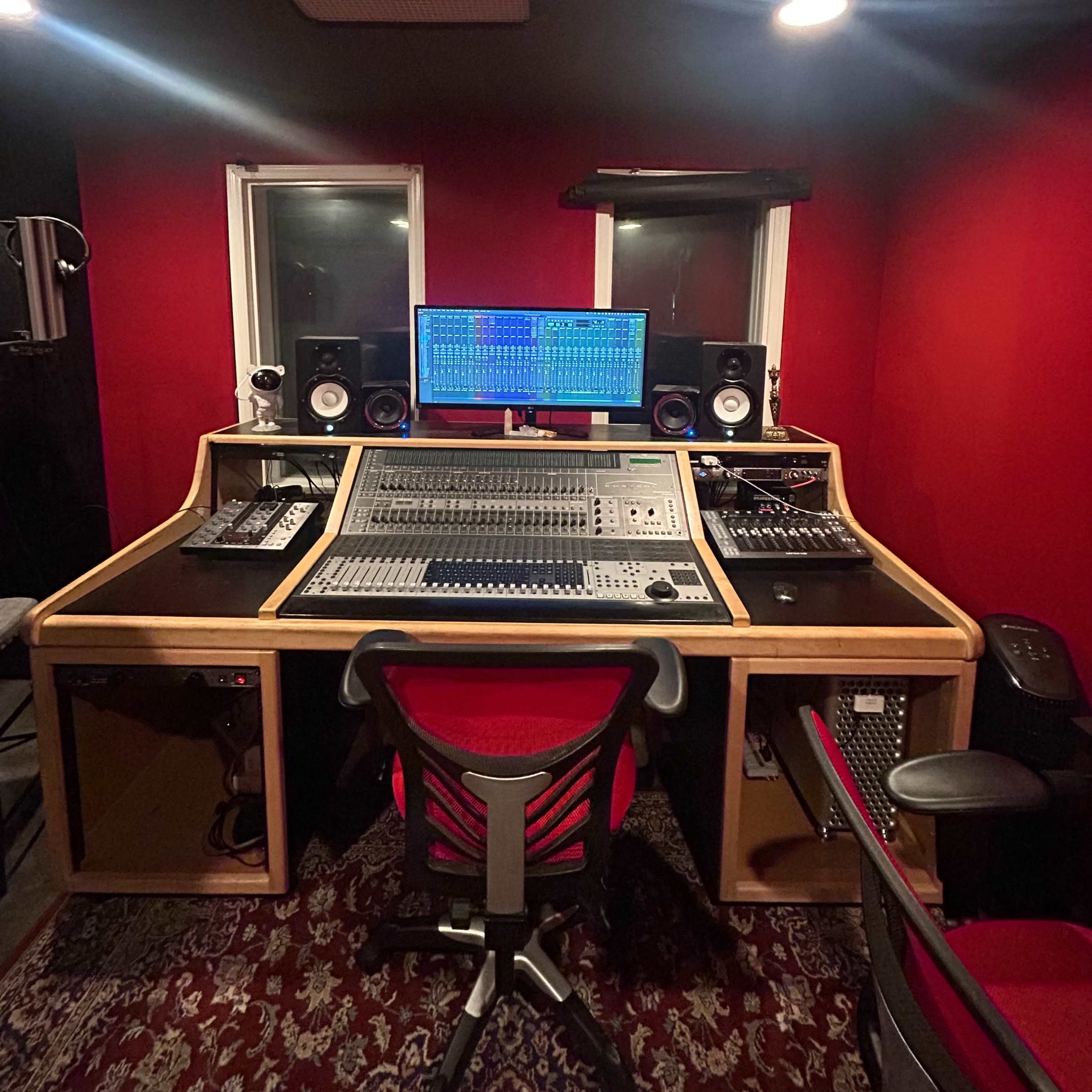 A recording studio features a large mixing console, computer monitor, and audio equipment set against red walls.
