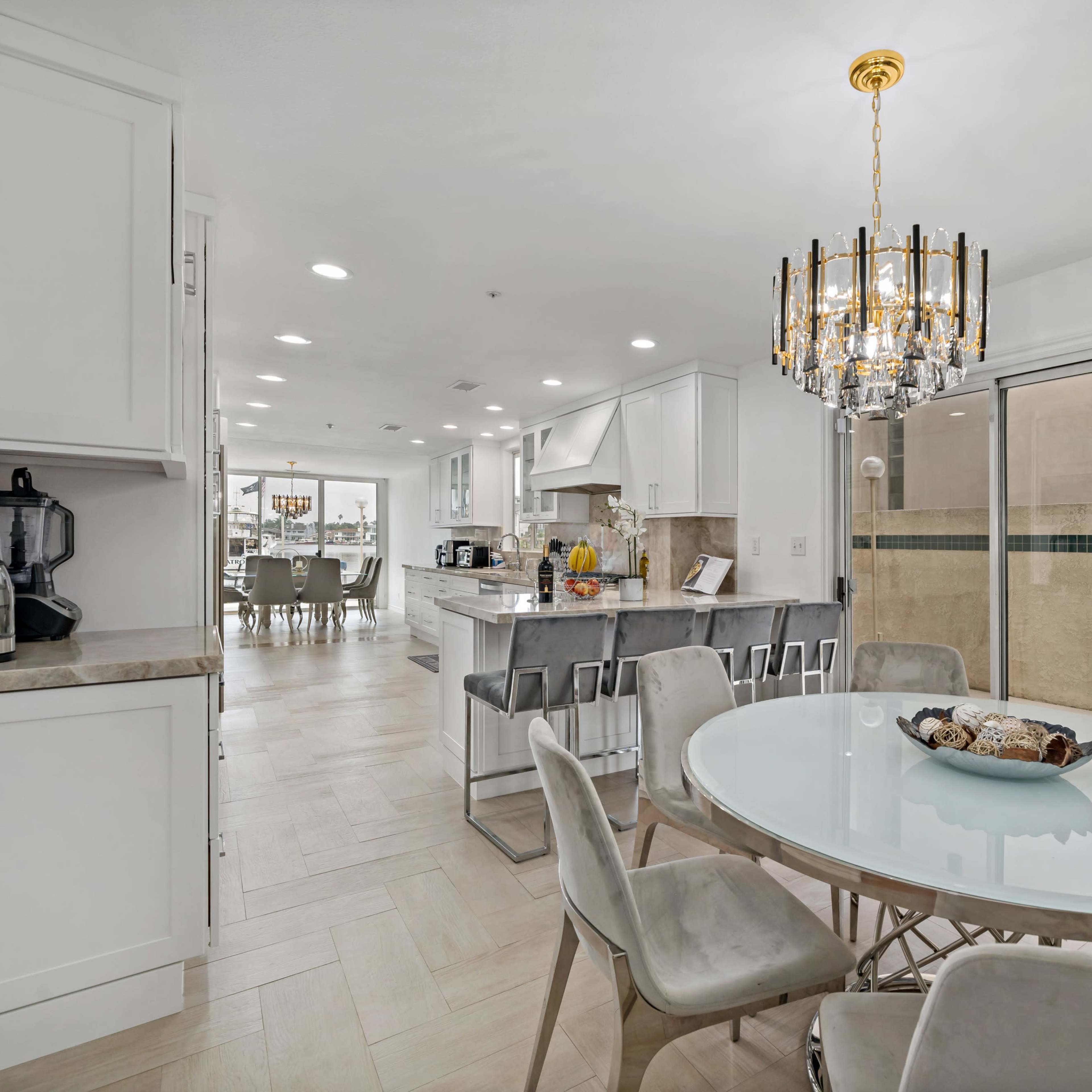 A modern kitchen and dining area with a round glass table surrounded by six chairs, illuminated by a chandelier.
