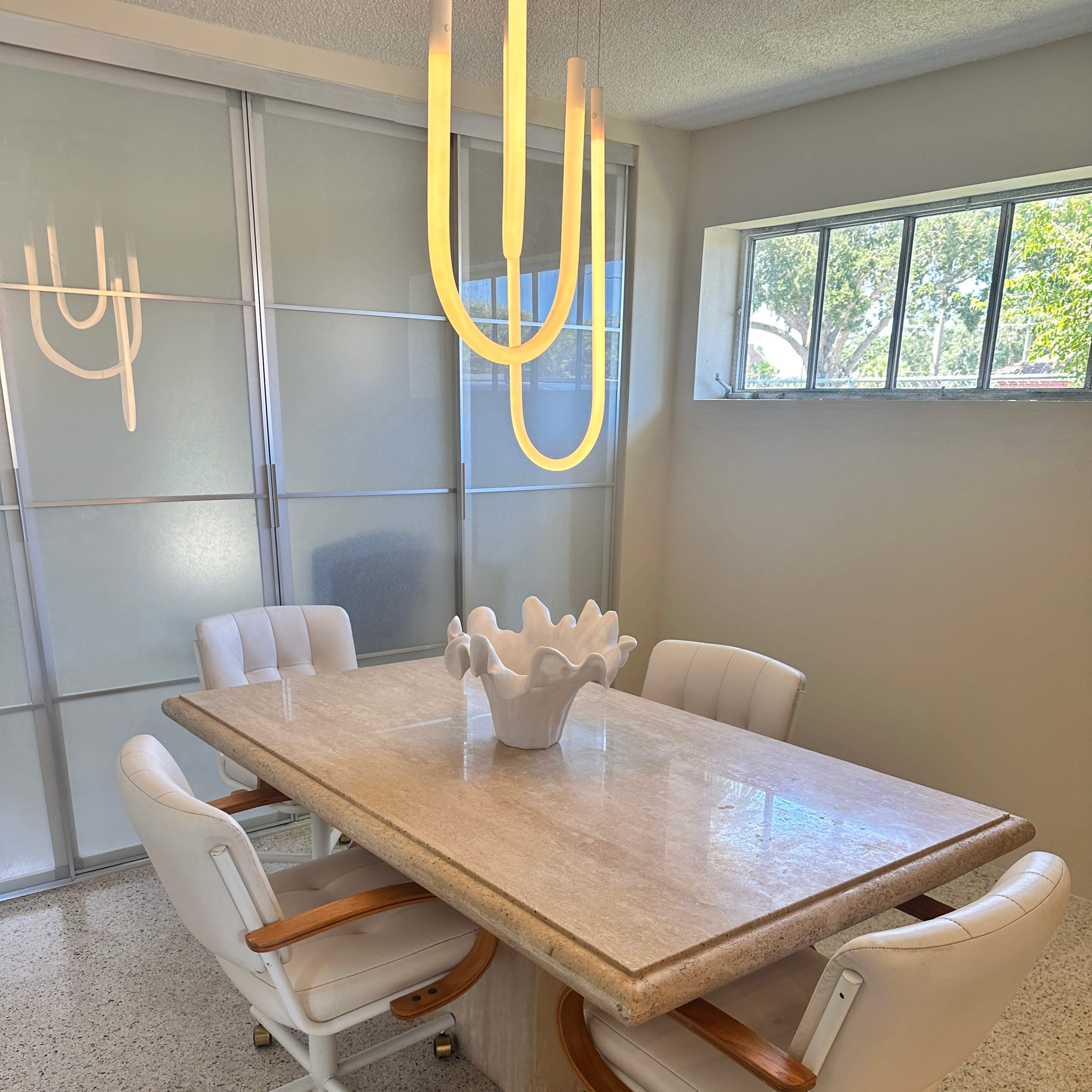A modern dining area features a rectangular stone table surrounded by four white chairs and illuminated by a contemporary hanging light fixture.