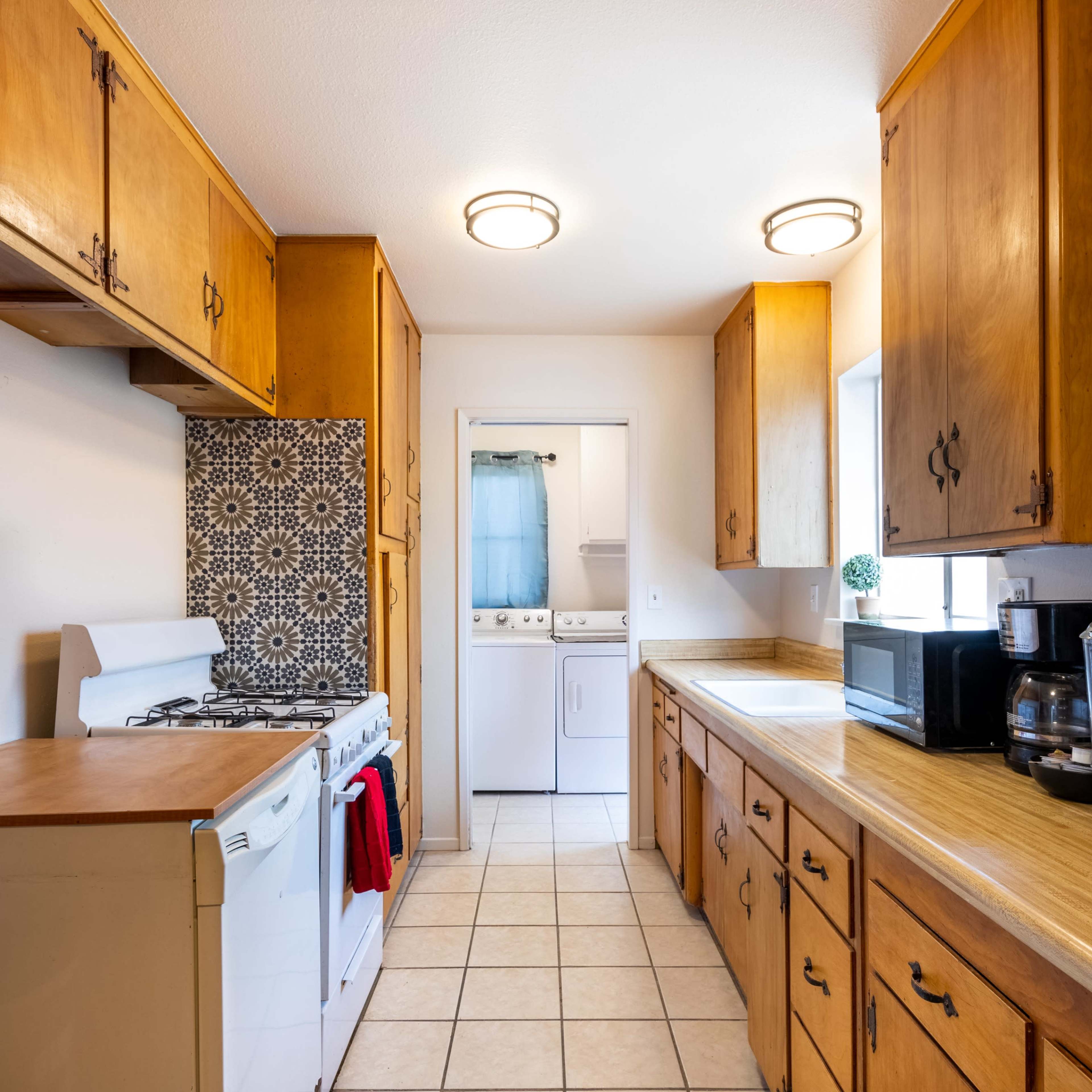 A narrow kitchen features wooden cabinets, a gas stove with a red towel, and a laundry area visible through a door at the end.