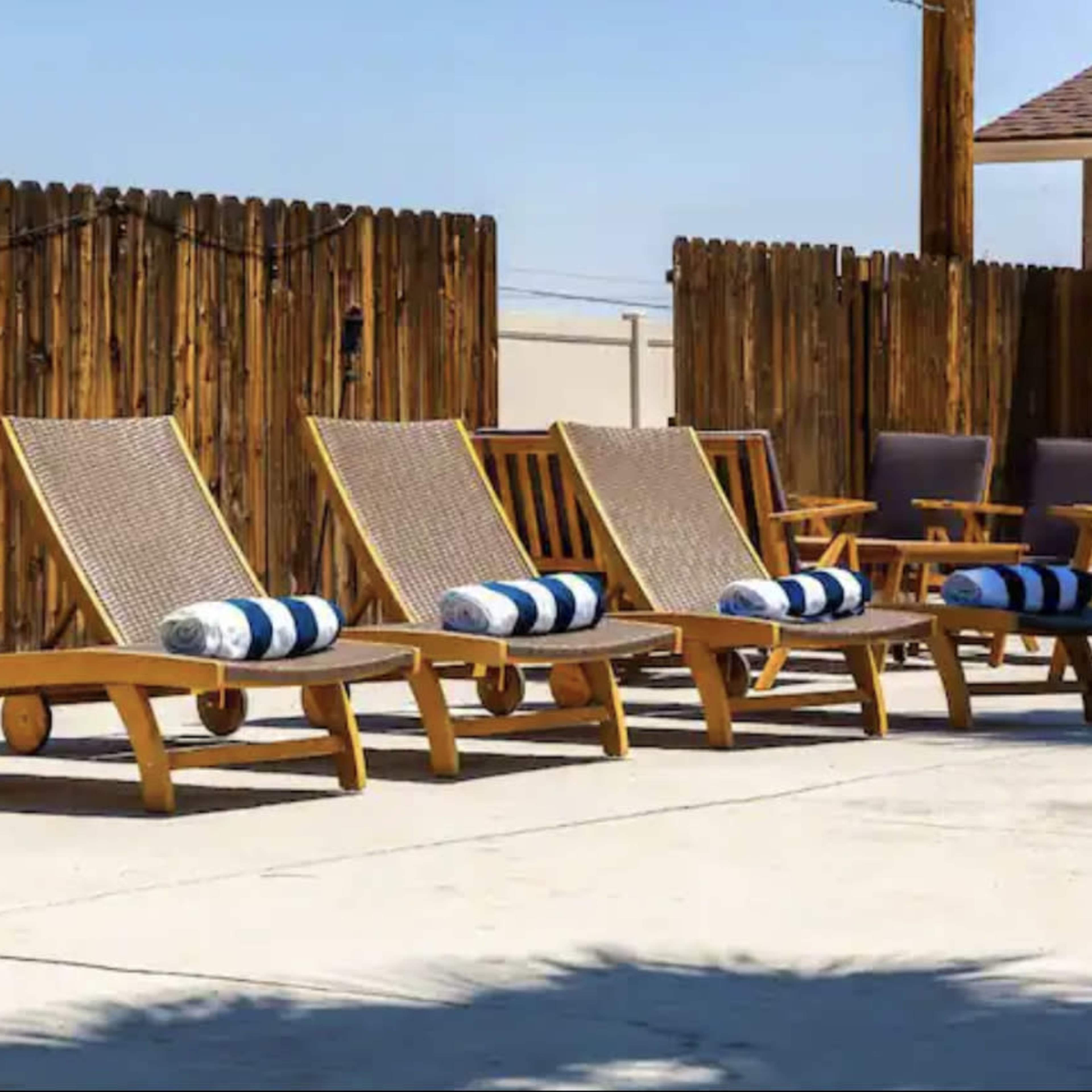 A row of wooden loungers with blue and white striped towels is arranged beside a concrete patio, in front of a wooden fence.