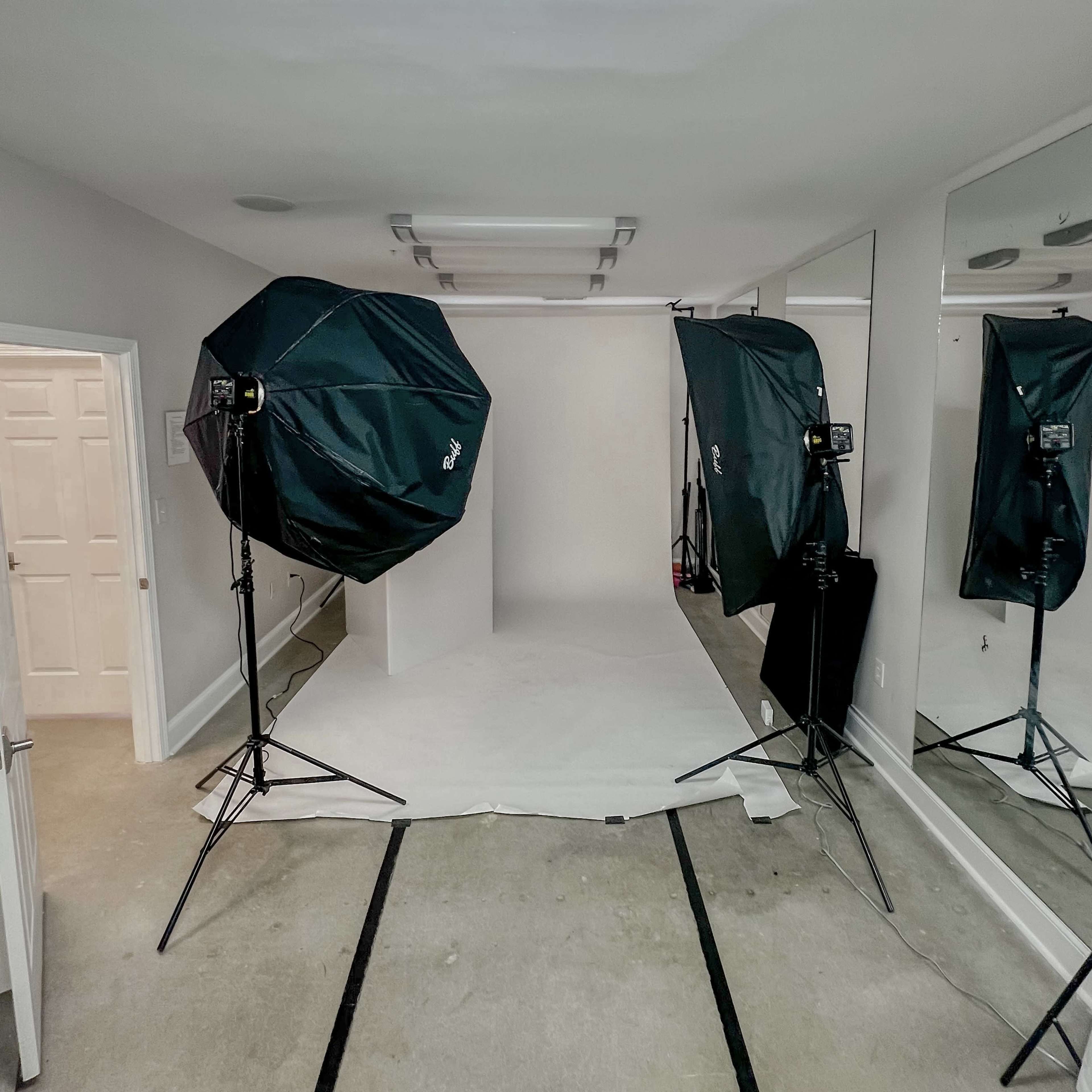 A photography studio equipped with two large softlight umbrellas facing a seamless backdrop, with mirrors and a white door visible in the background.