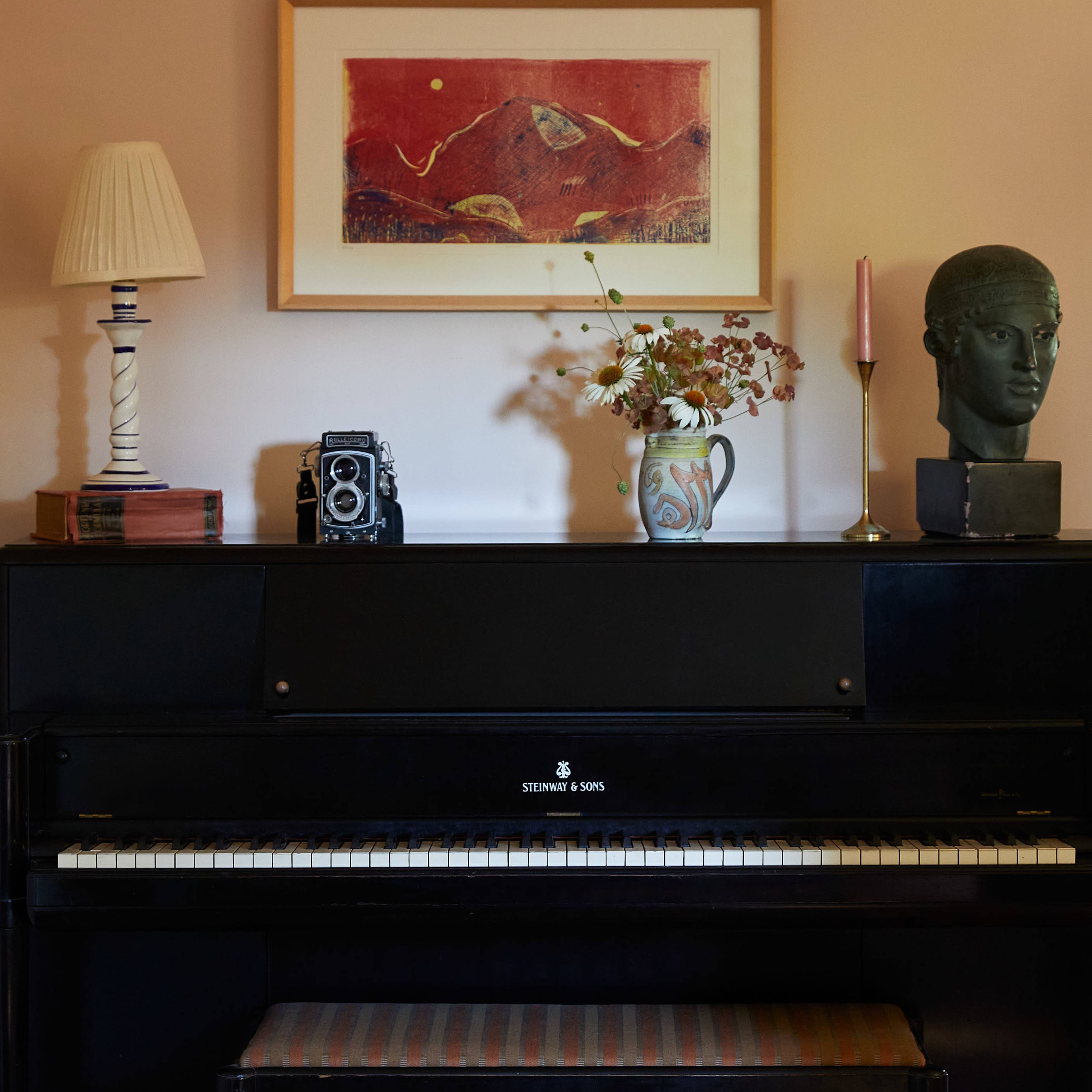A black piano with sheet music sits below a framed artwork, accompanied by a vintage camera, a vase of dried flowers, a candle holder, and a bust on the shelf above.