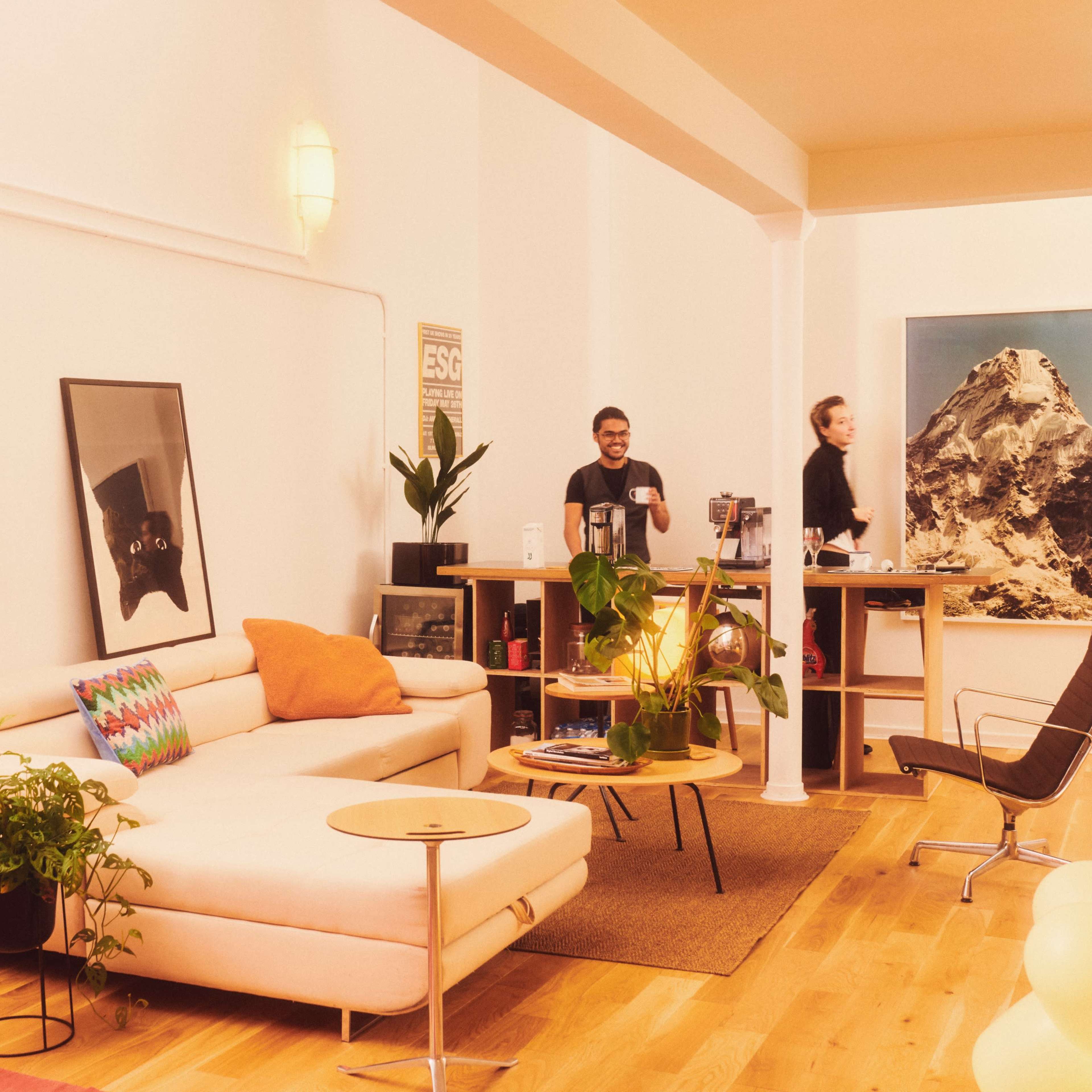 A modern office space featuring a light-colored couch, a wooden desk with two people, and a large mountain artwork on the wall.