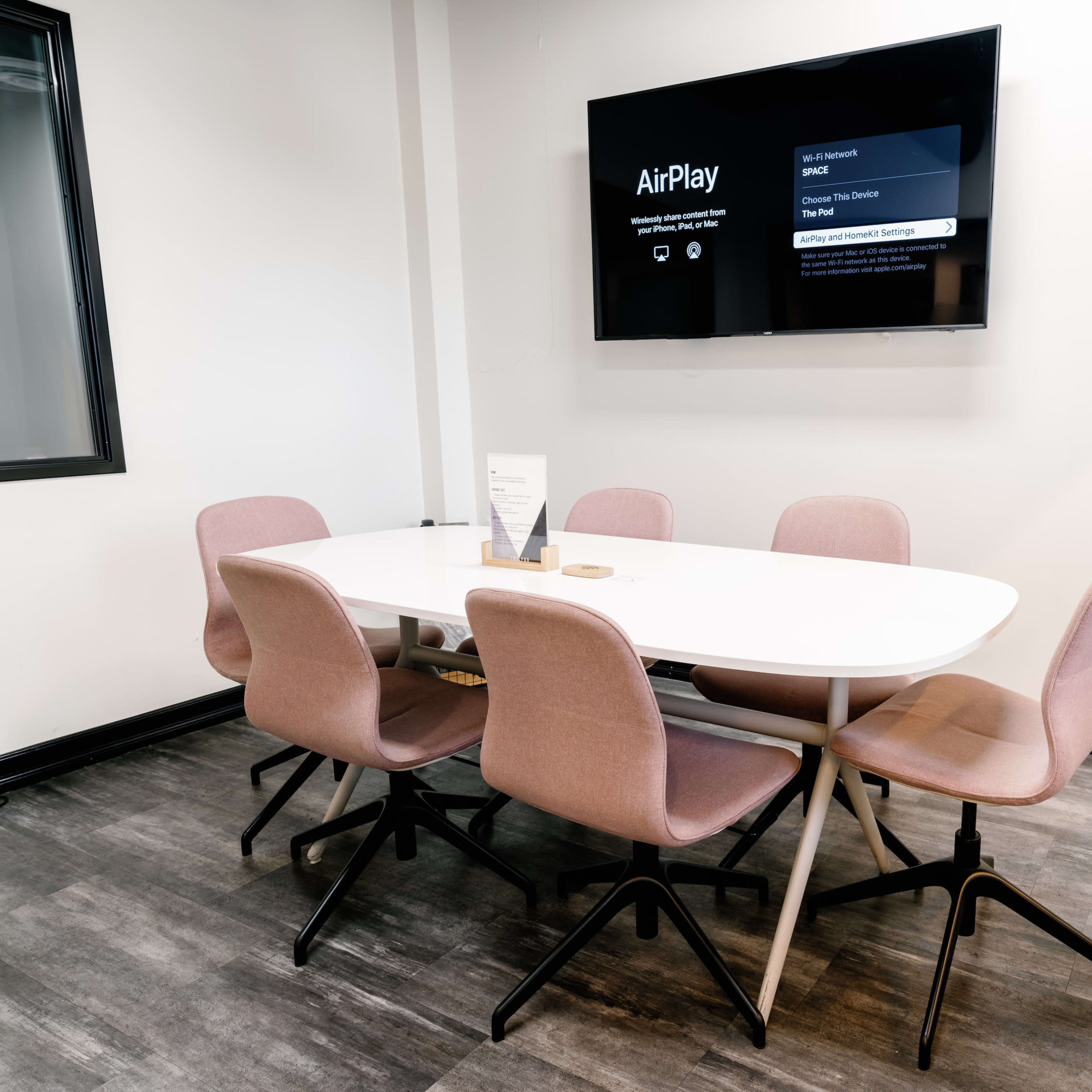 A modern conference room features a white oval table surrounded by six pink chairs, with a wall-mounted screen displaying an AirPlay menu.