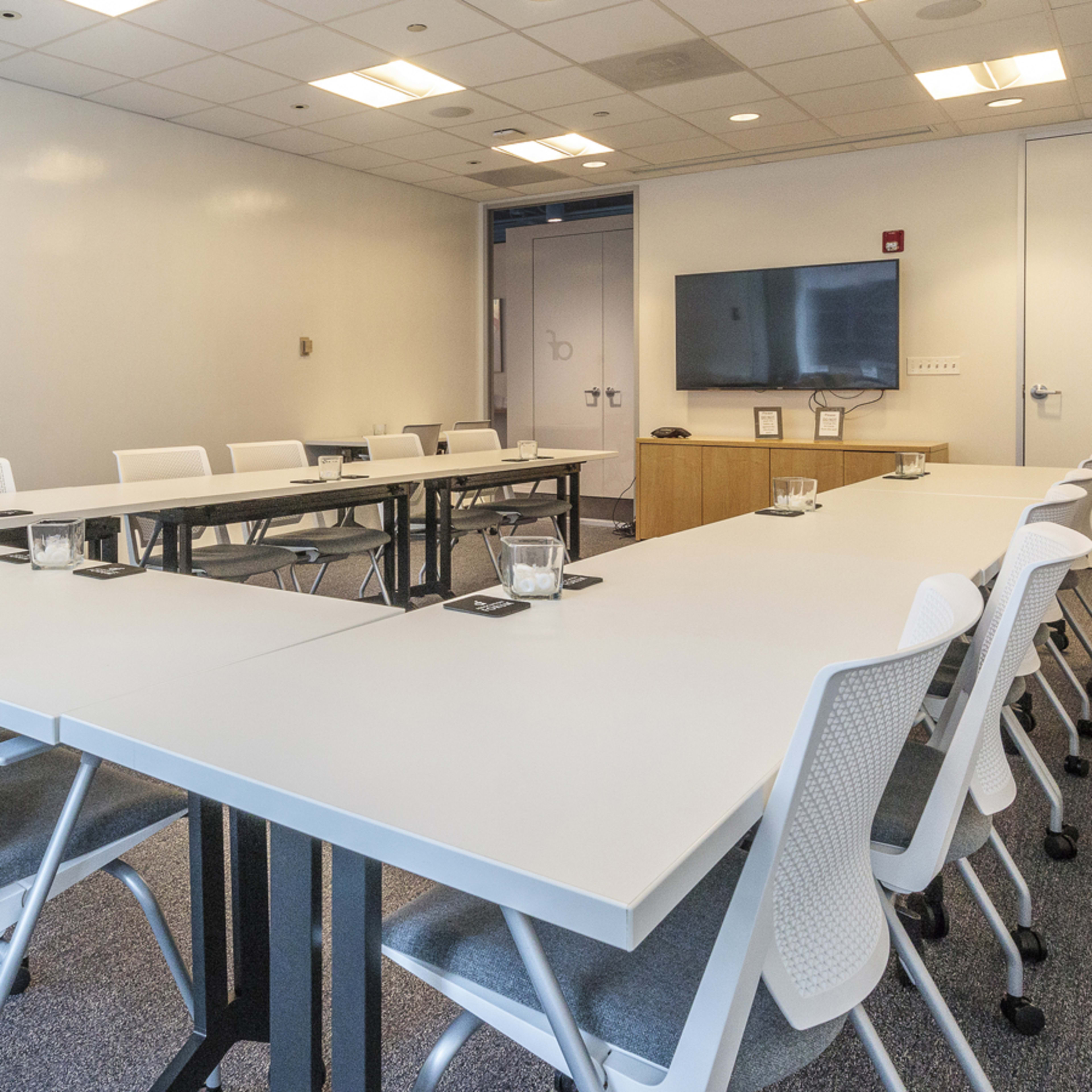 A modern conference room features multiple tables arranged in a U-shape, with chairs, a television mounted on the wall, and a glass panel door.