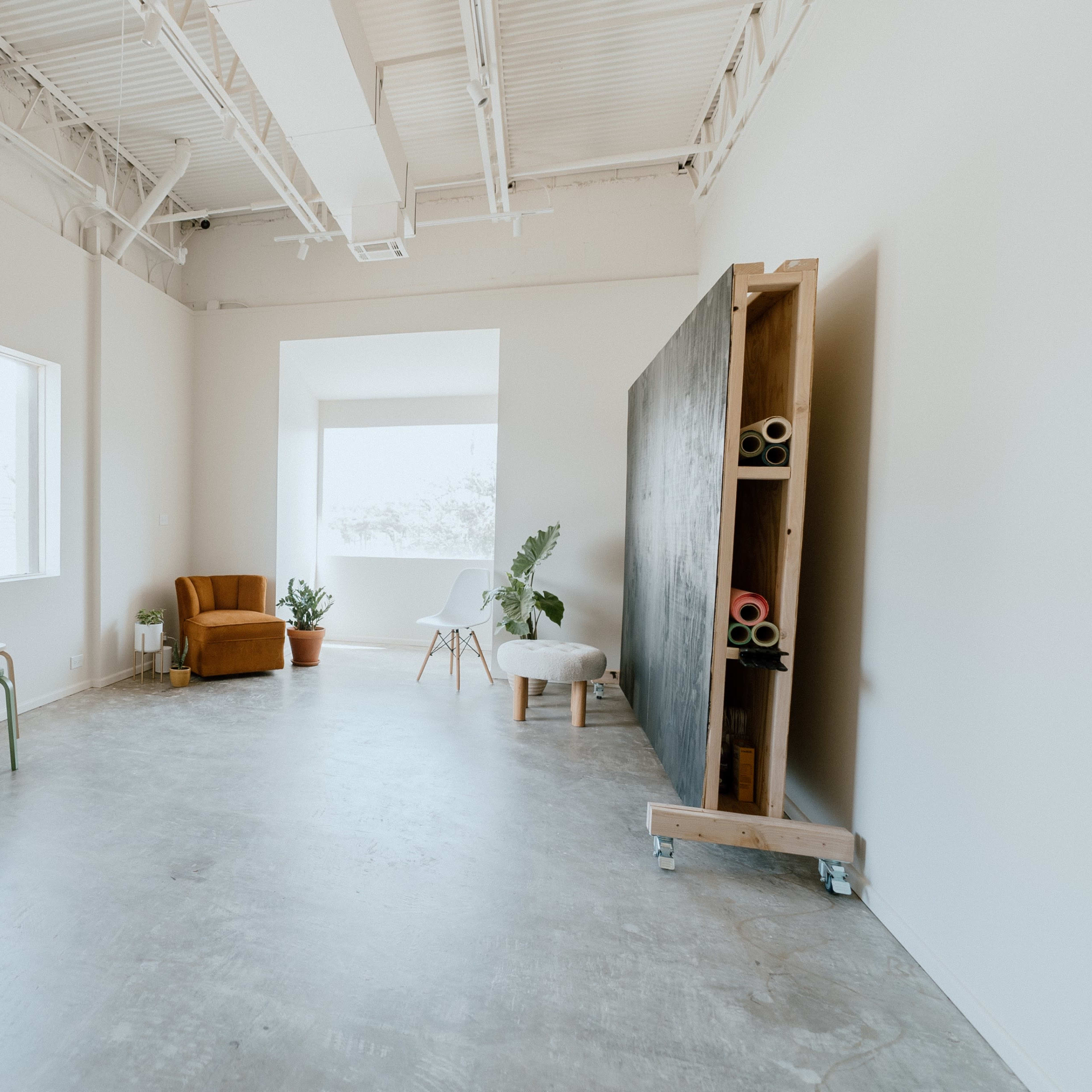 A spacious, minimalist room with a large window, a brown armchair, a small table, a plant, and a display of rolled materials against a wall.