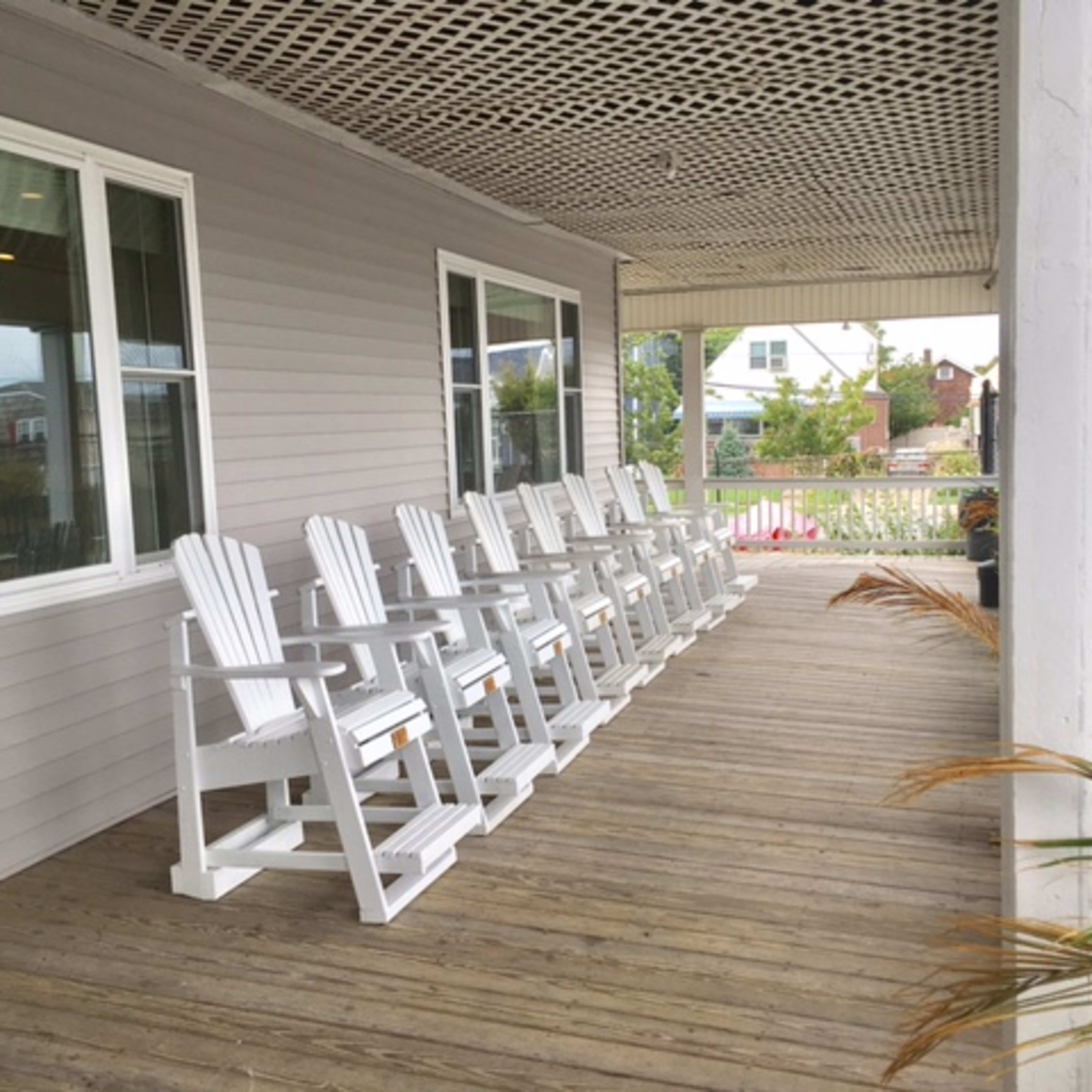 A row of white Adirondack chairs is lined up on a wooden porch with a slatted ceiling.