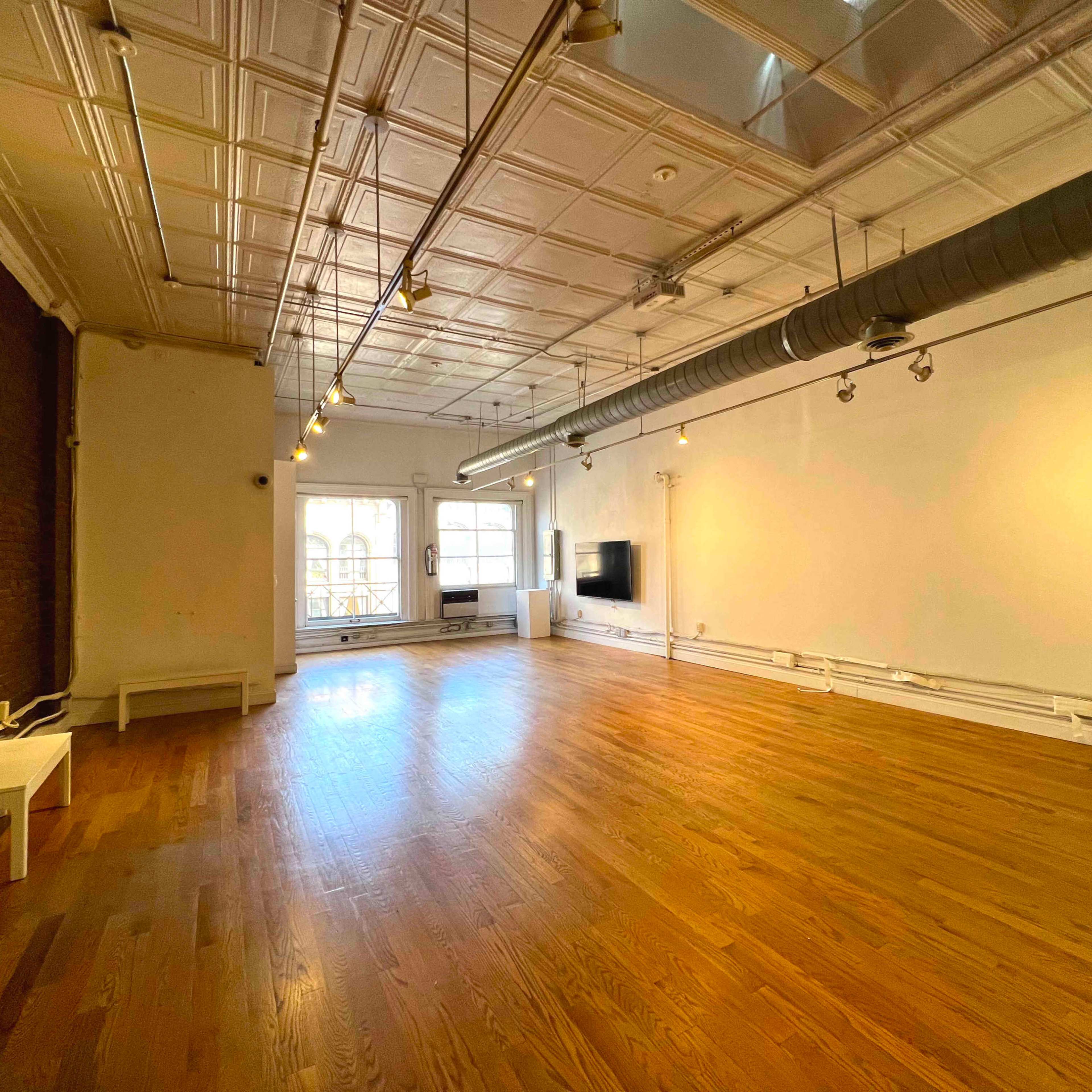 A spacious, empty room with hardwood floors, exposed brick walls, and large windows letting in natural light.