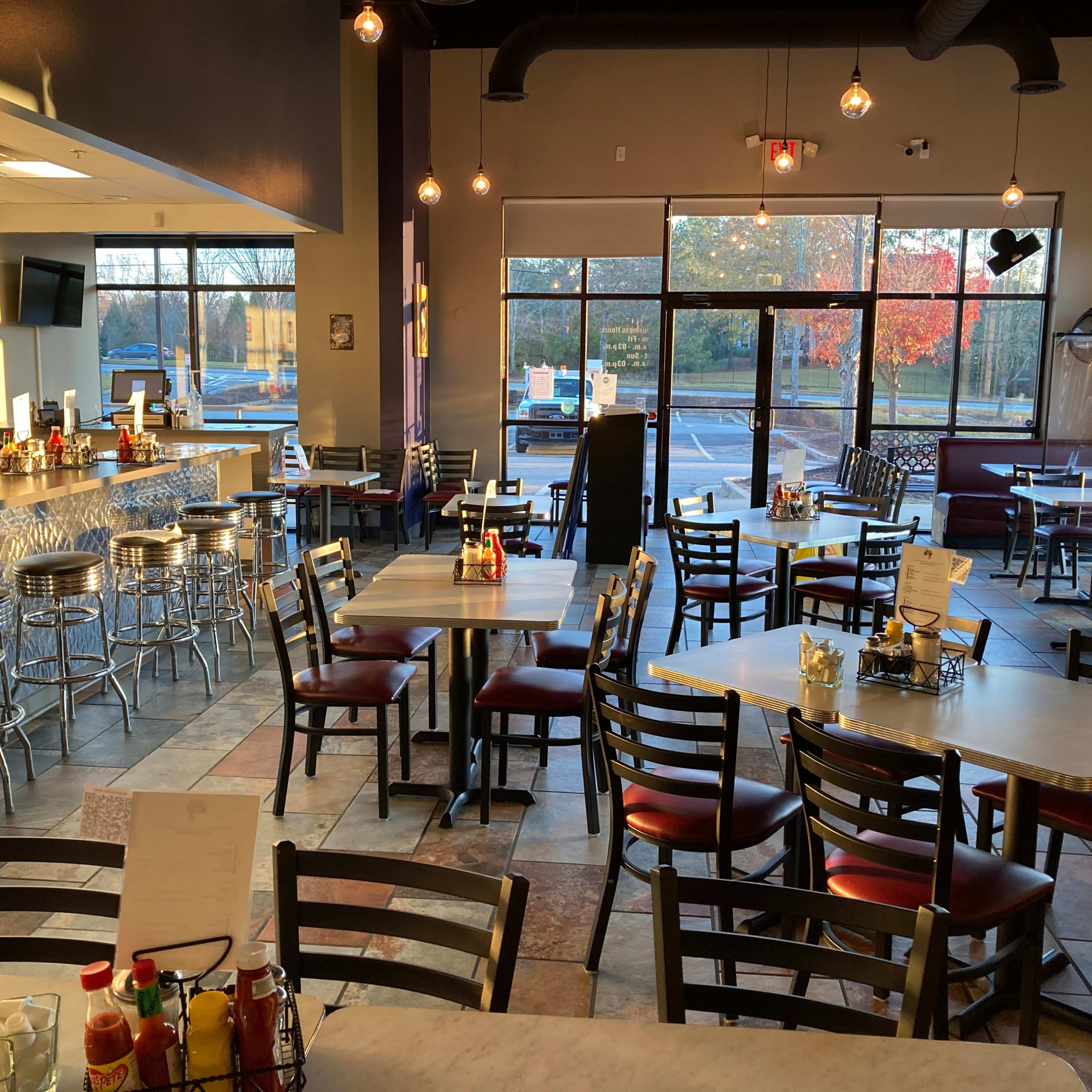 A modern restaurant interior with empty tables and chairs, a counter area, and large windows displaying an outdoor view.