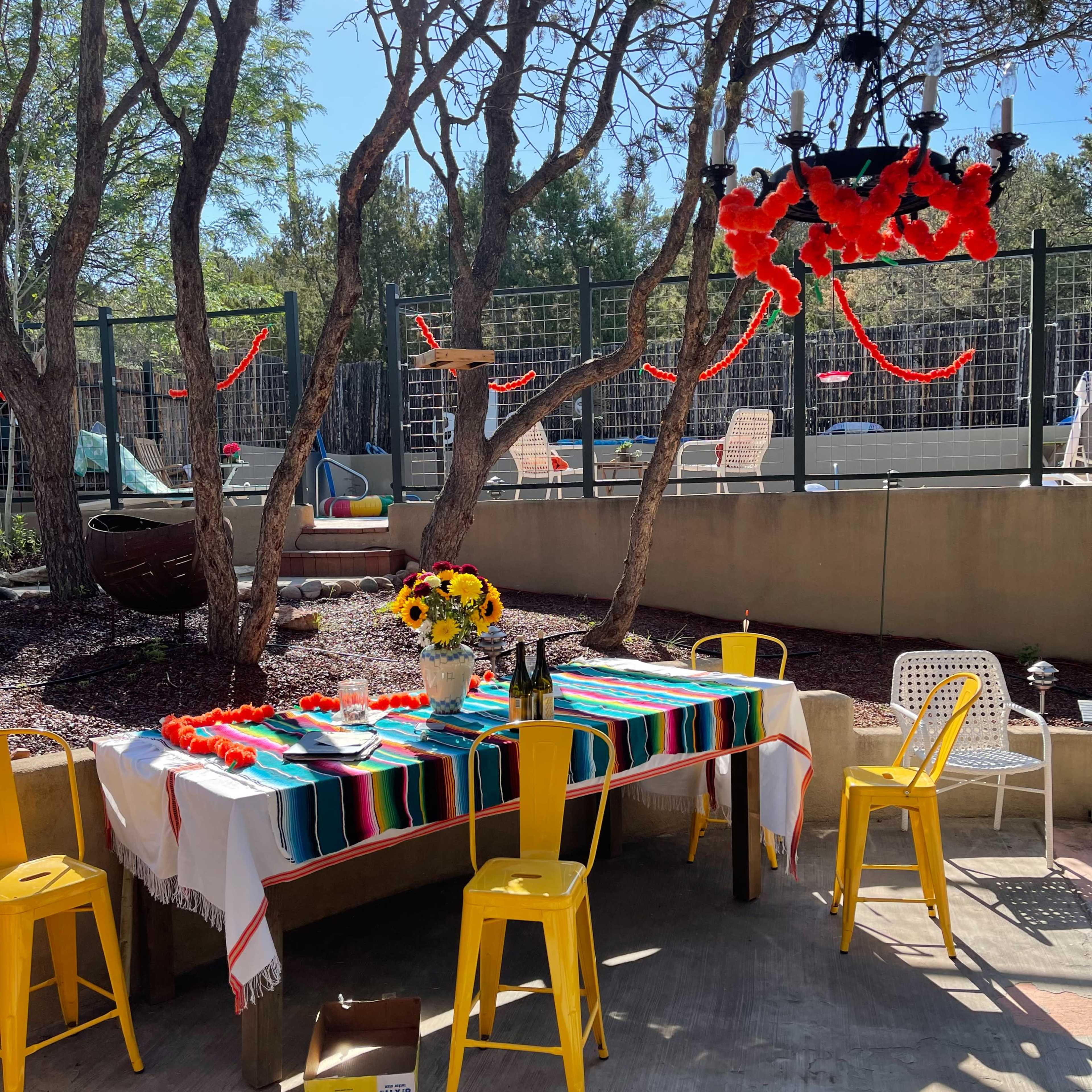 A table covered with a colorful striped cloth is set outside under trees, surrounded by bright yellow chairs and decorated with sunflowers and red streamers.