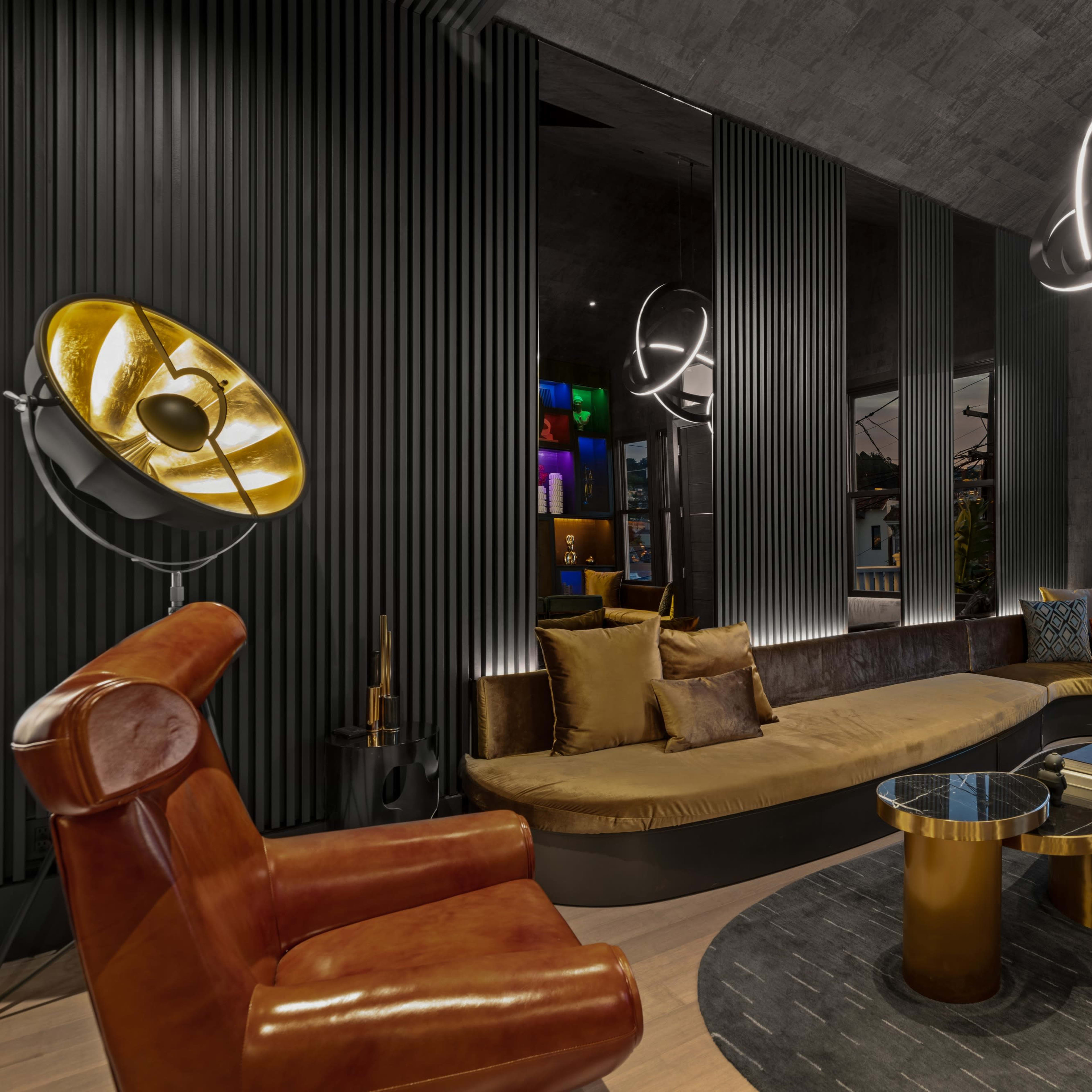 A modern lounge area featuring a circular sofa, a round table, a leather armchair, and stylish light fixtures against a backdrop of textured walls and large windows.