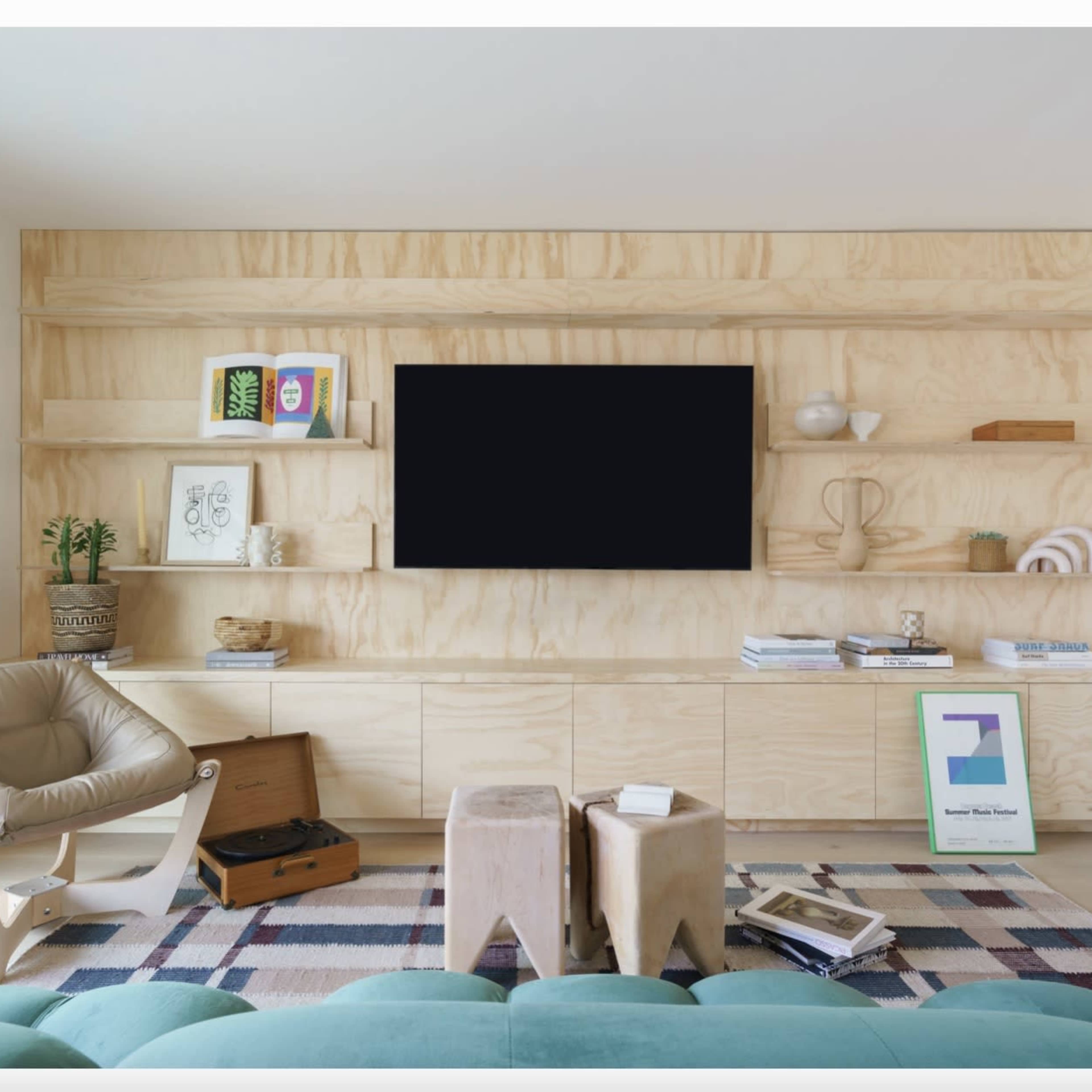 A modern living room features a plywood wall unit with a large black screen and minimalist decor, complemented by a light-colored armchair and a patterned rug.