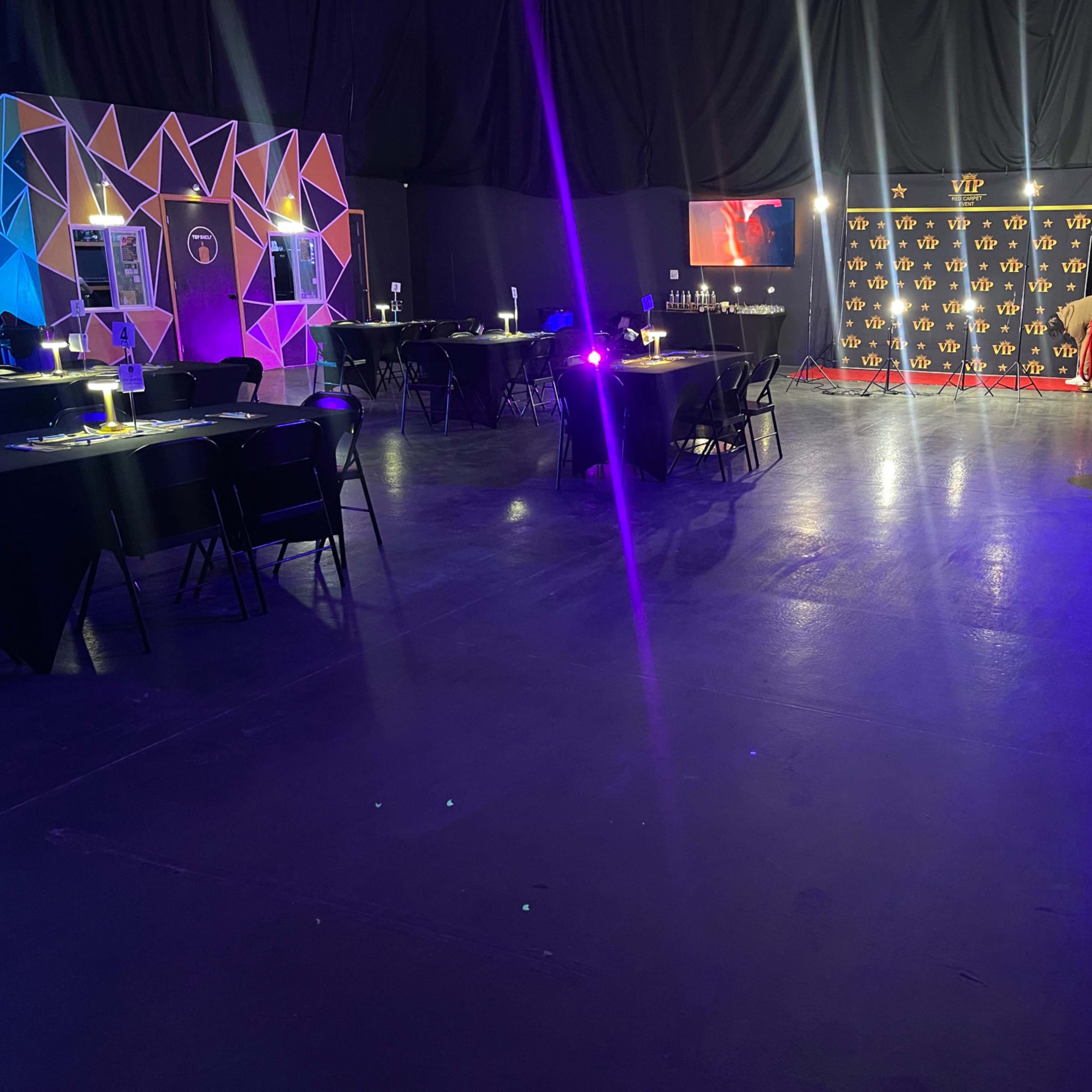 An event space with black tables and chairs, colorful geometric decorations on one wall, and a lit backdrop with a logo in the distance.