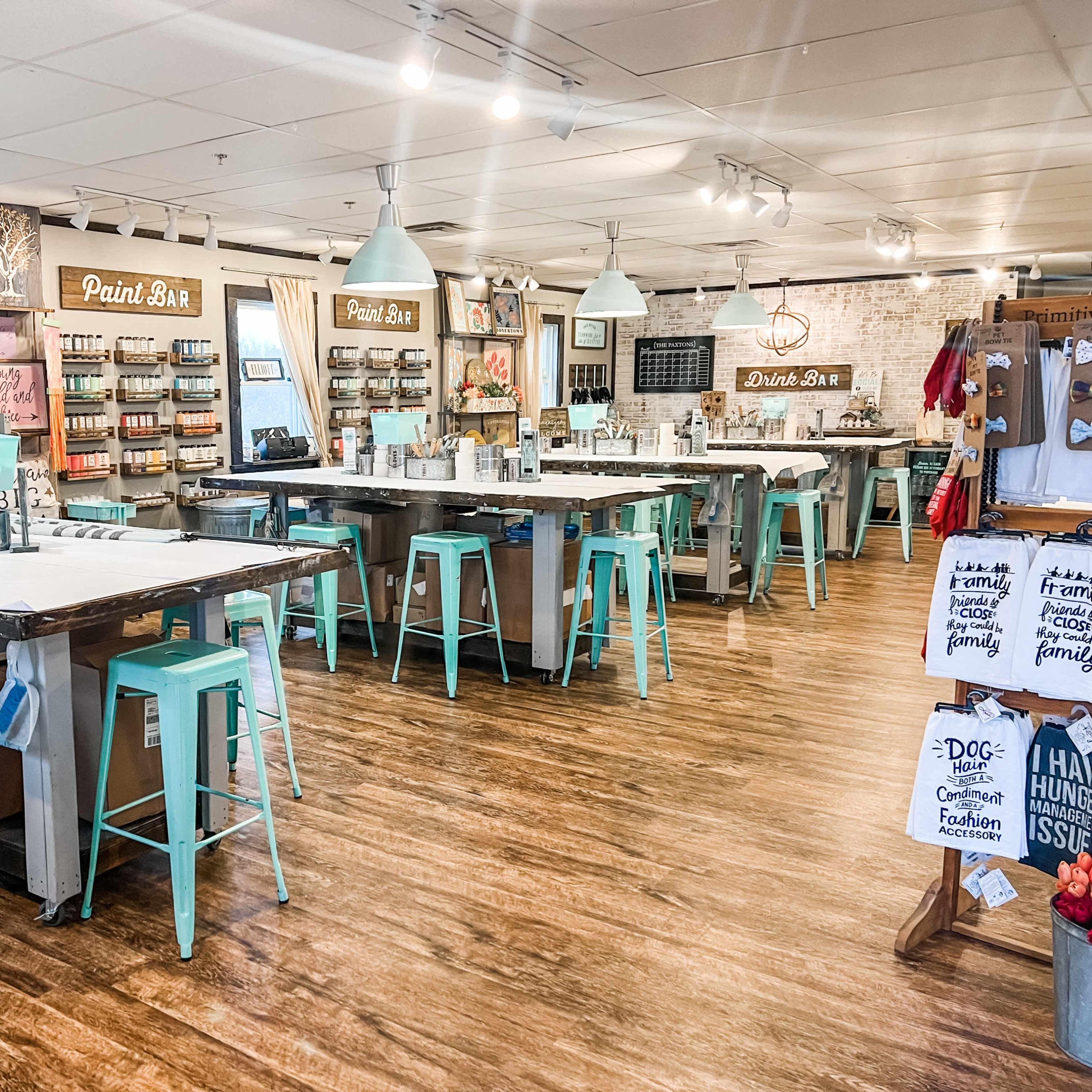 A brightly-lit craft studio featuring multiple tables with mint green stools, a paint bar, and shelves stocked with various paint supplies.