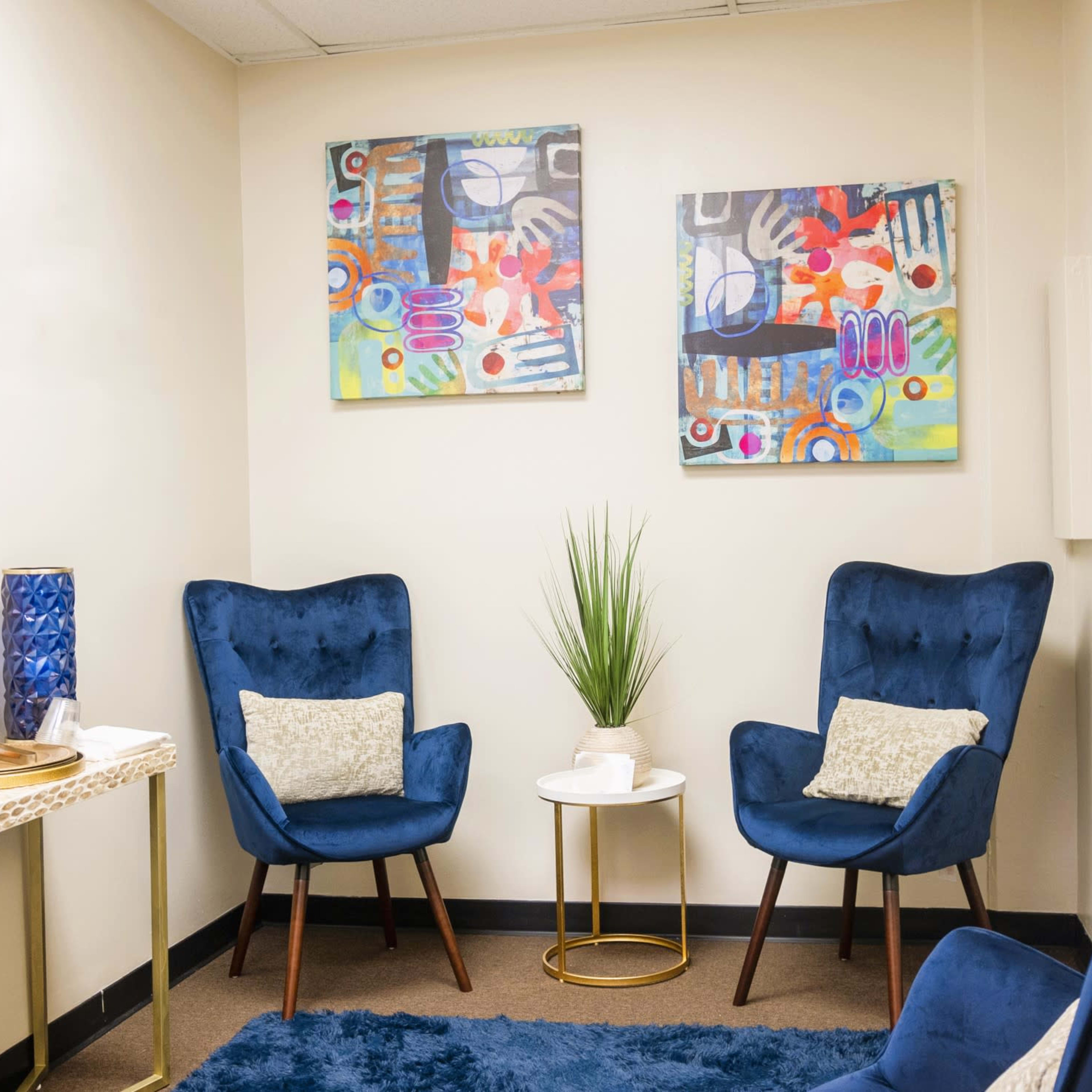 A small, neatly arranged room with two blue armchairs, a round table, and colorful artwork on the walls.