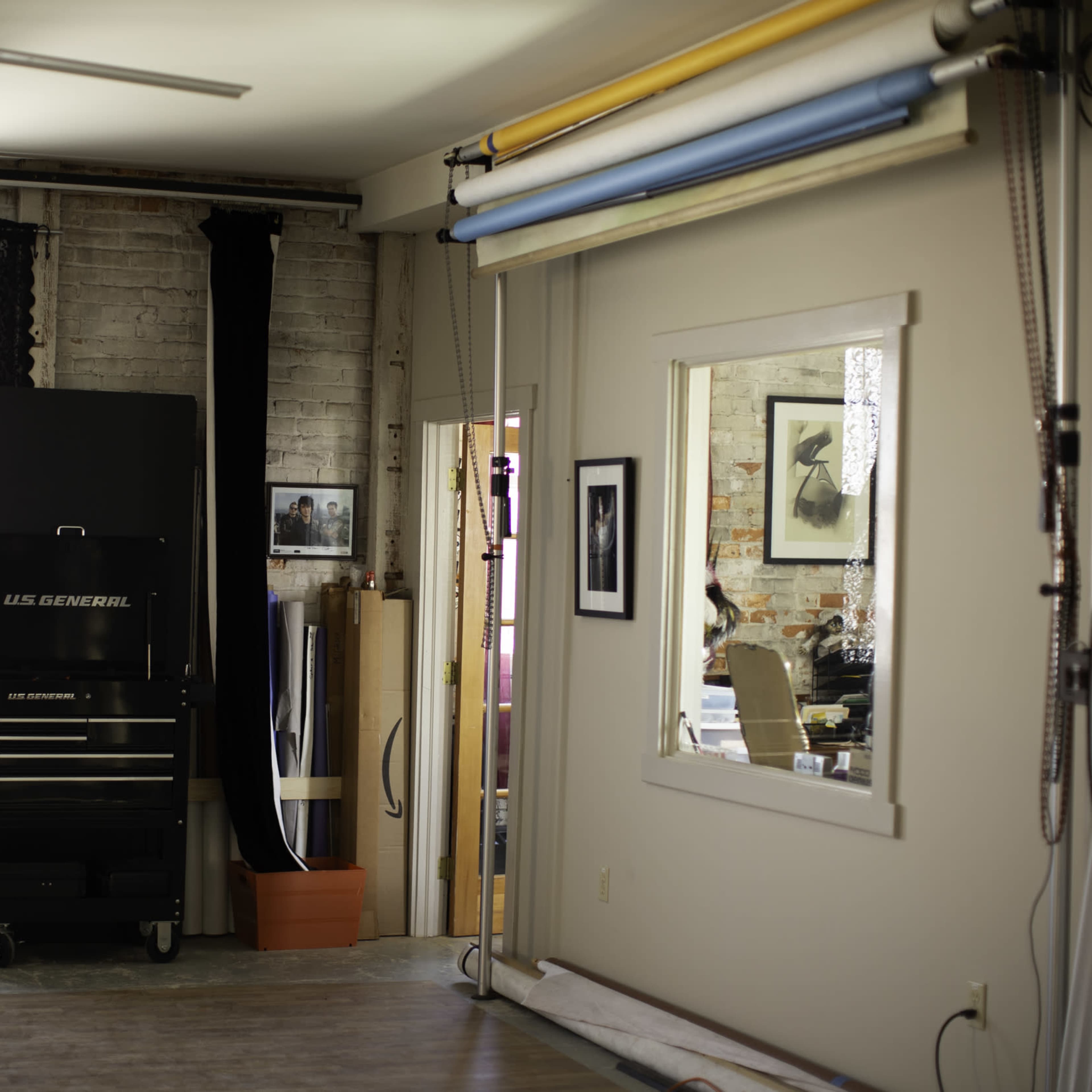 A well-lit studio space with a large window, a tool cabinet, a mirror, and different colored backdrop rolls hanging on the wall.