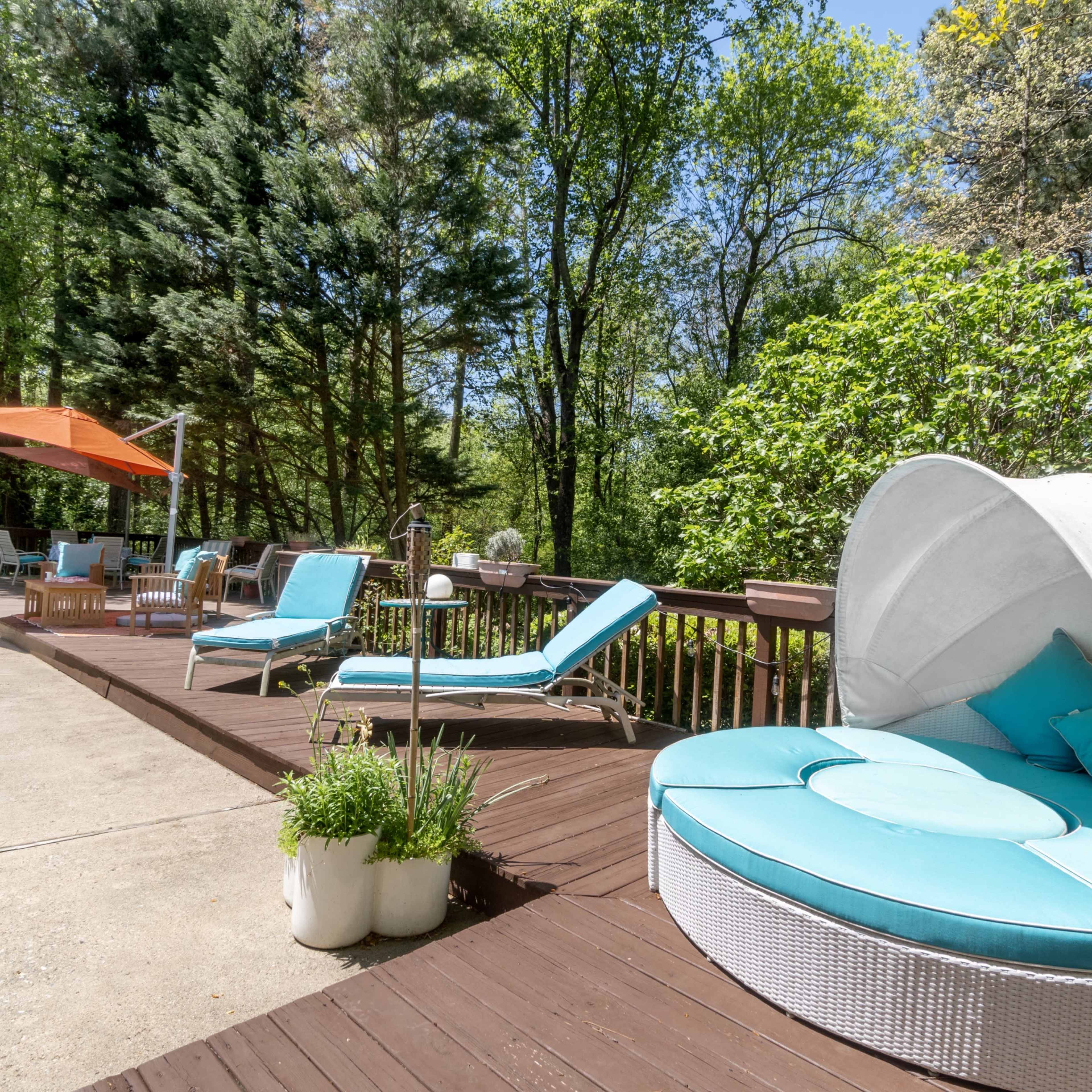 A poolside area featuring a round, cushioned lounge chair, two chaise lounges, and an orange umbrella, surrounded by lush greenery.
