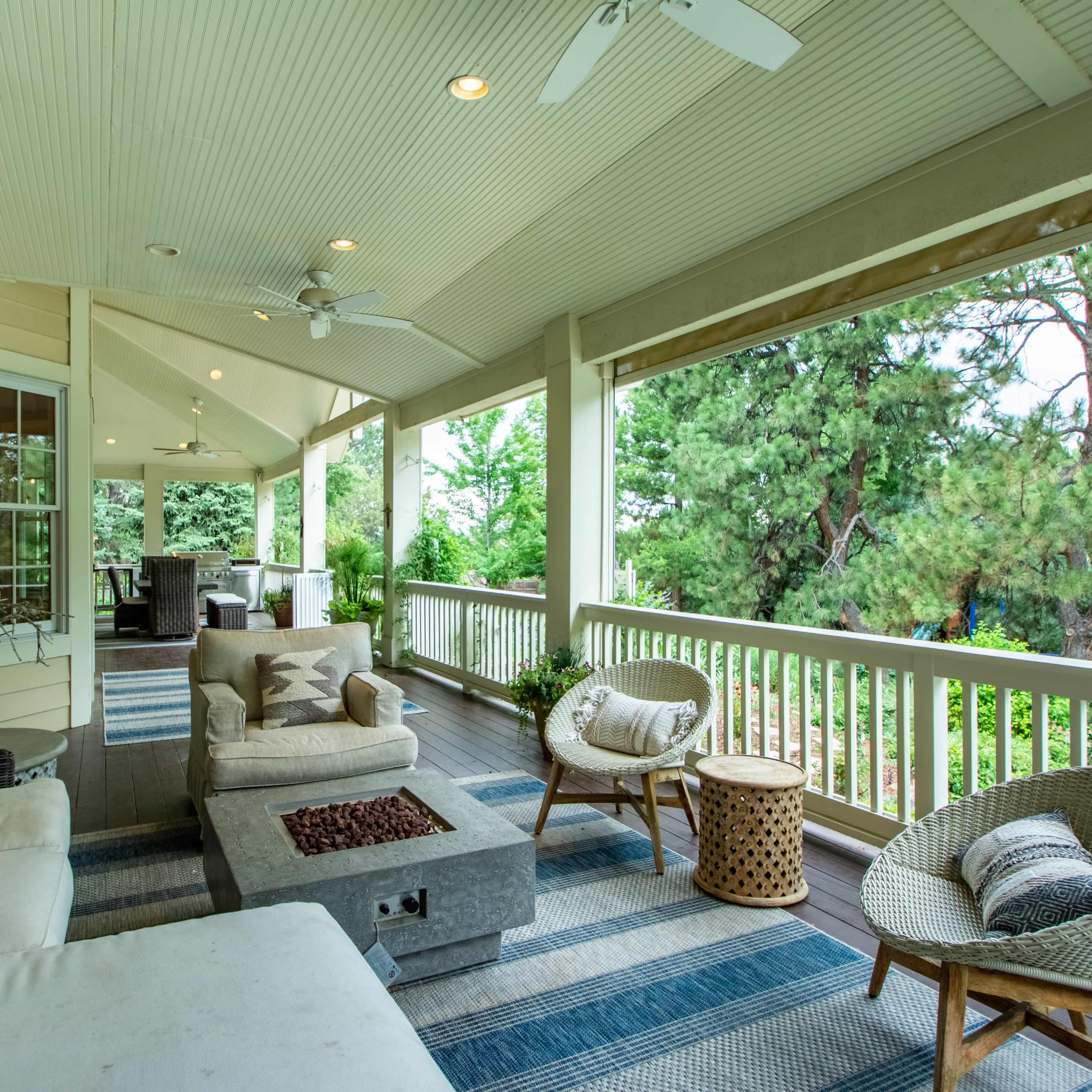 A covered porch features a seating area with a sectional couch, coffee table, and two chairs, surrounded by greenery and trees.