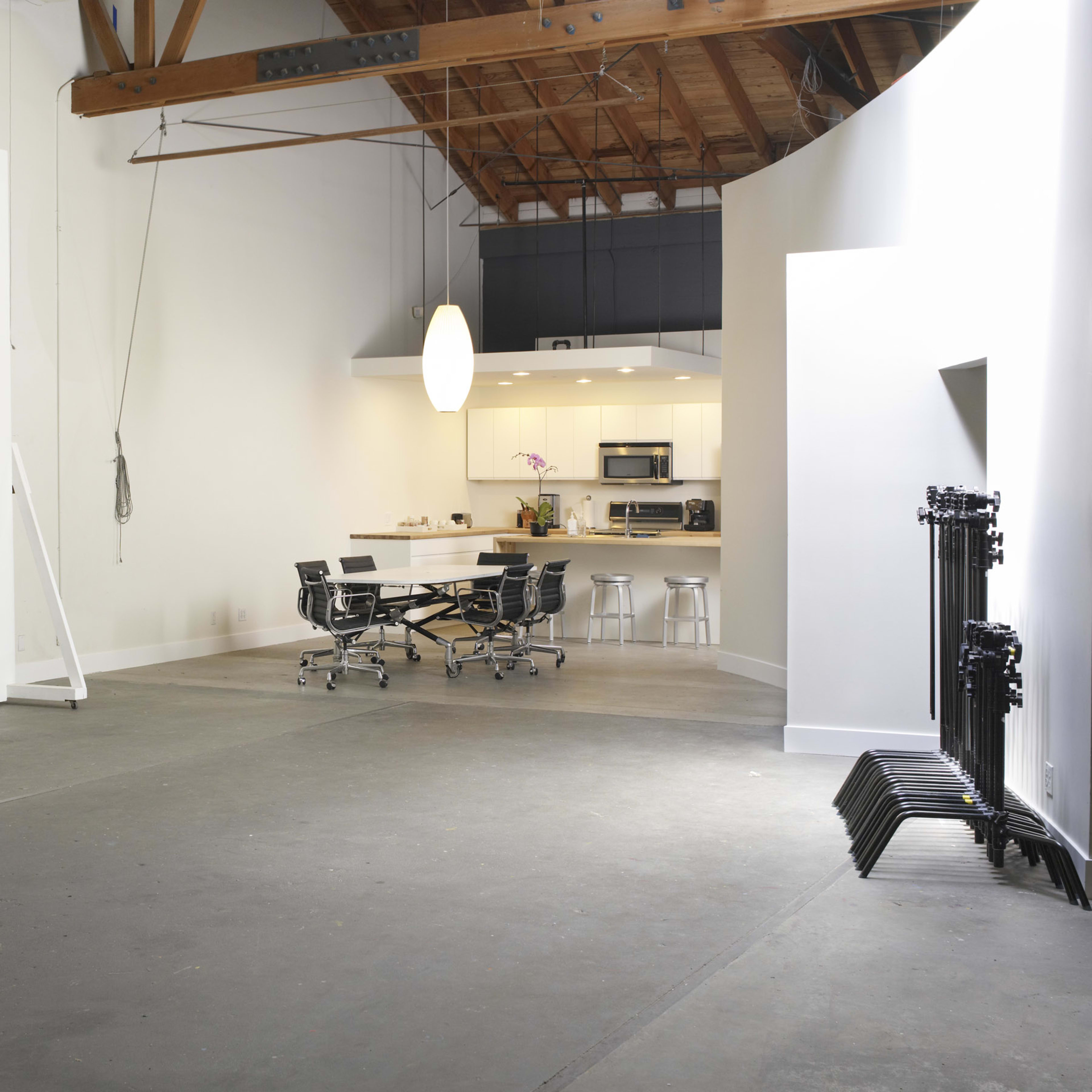 A spacious studio with a large white wall, a kitchen area in the background, and a meeting table surrounded by chairs.