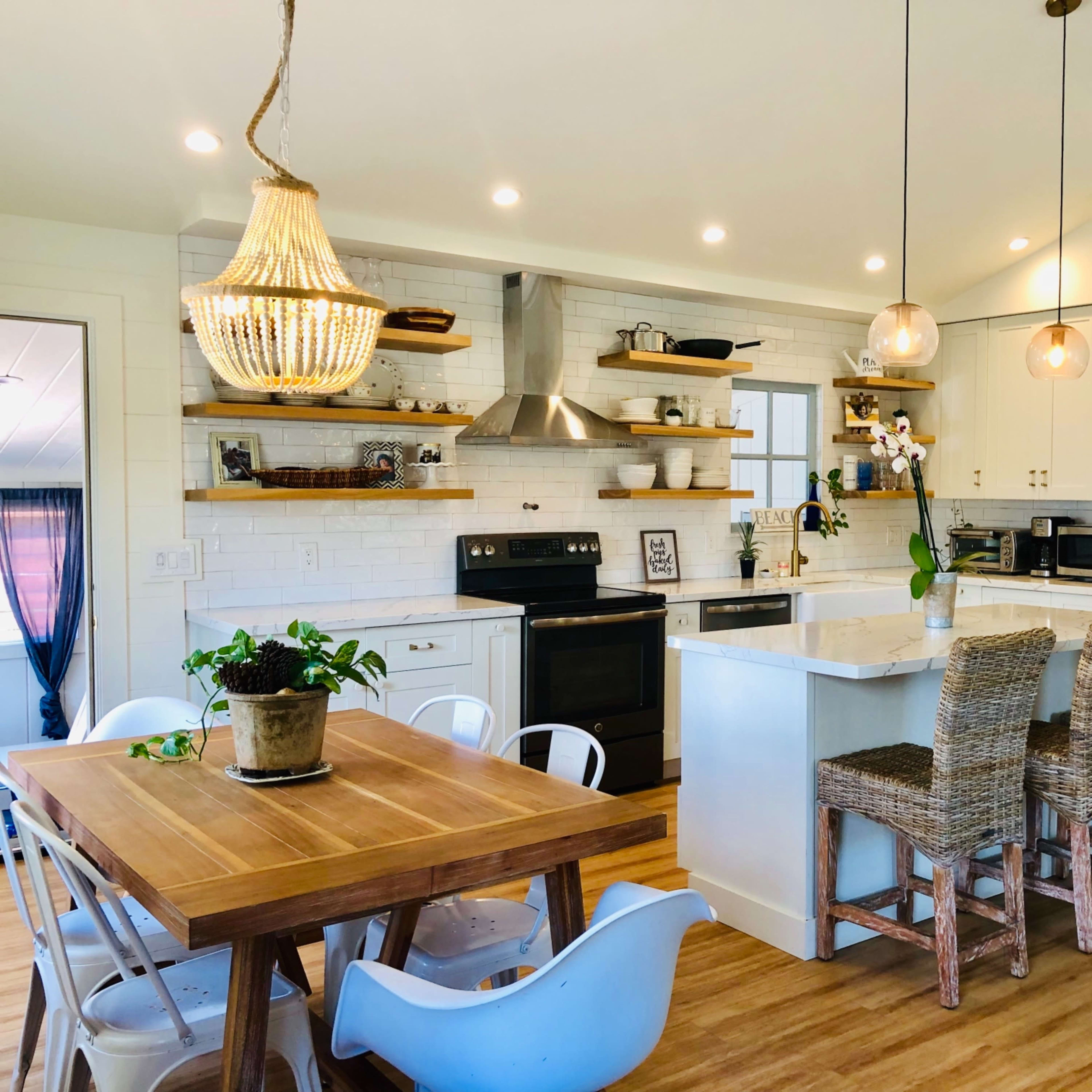 A modern kitchen features white cabinetry, open shelving with decorative items, a large island with seating, and a wooden dining table.