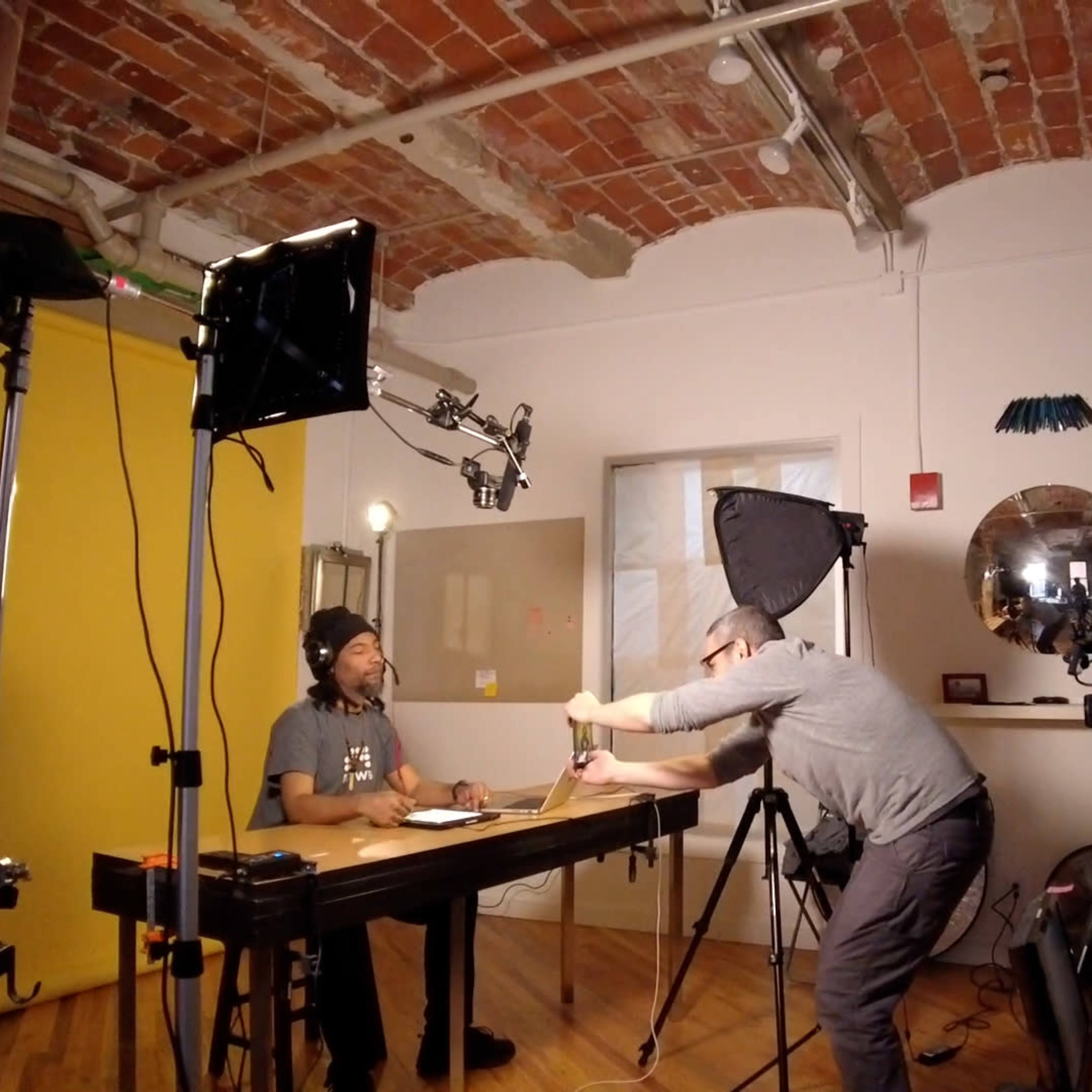 A video production setup features a man sitting at a table, while a crew member adjusts a light near a camera on a tripod.