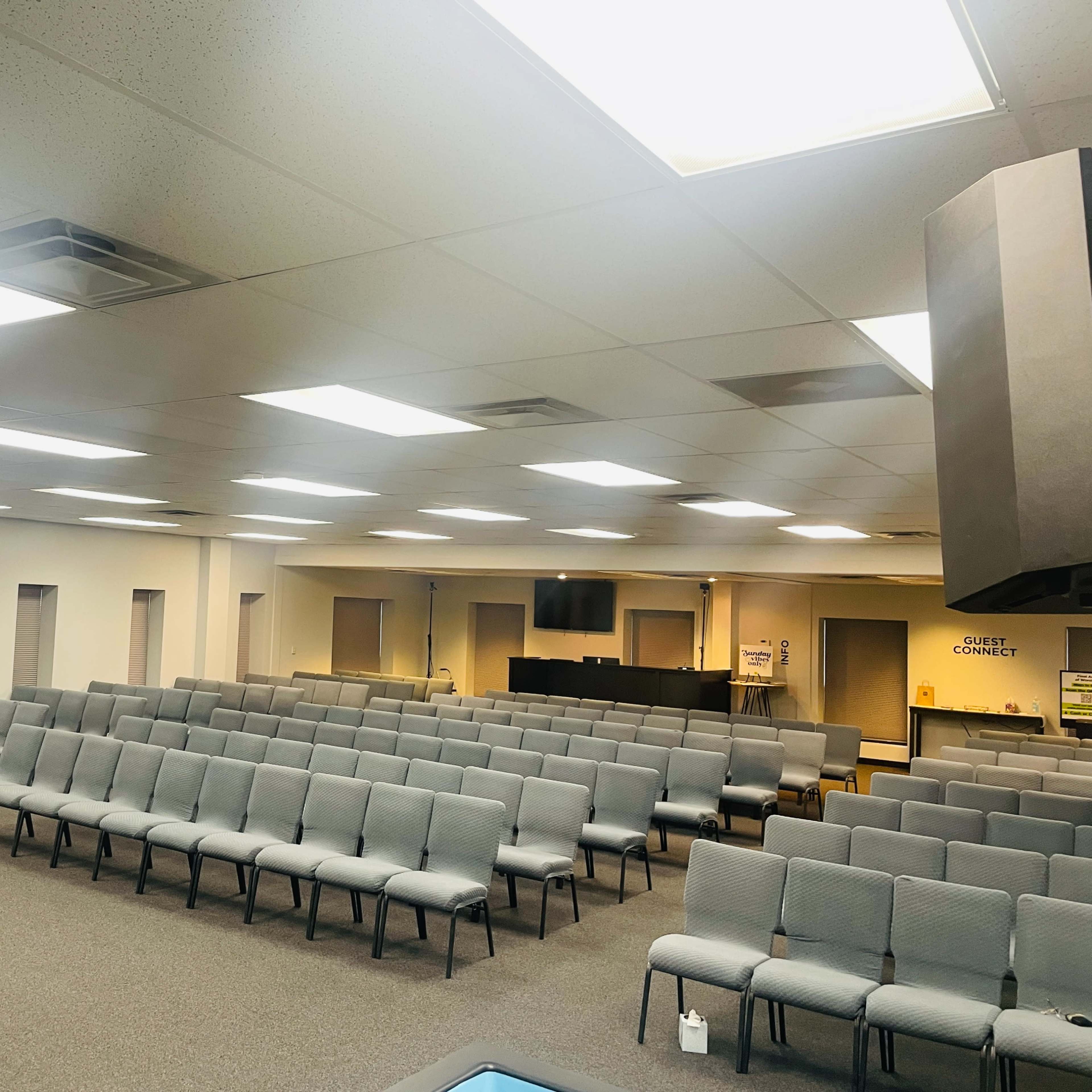 An empty, well-lit auditorium with rows of gray chairs facing a podium and a large screen on one side.