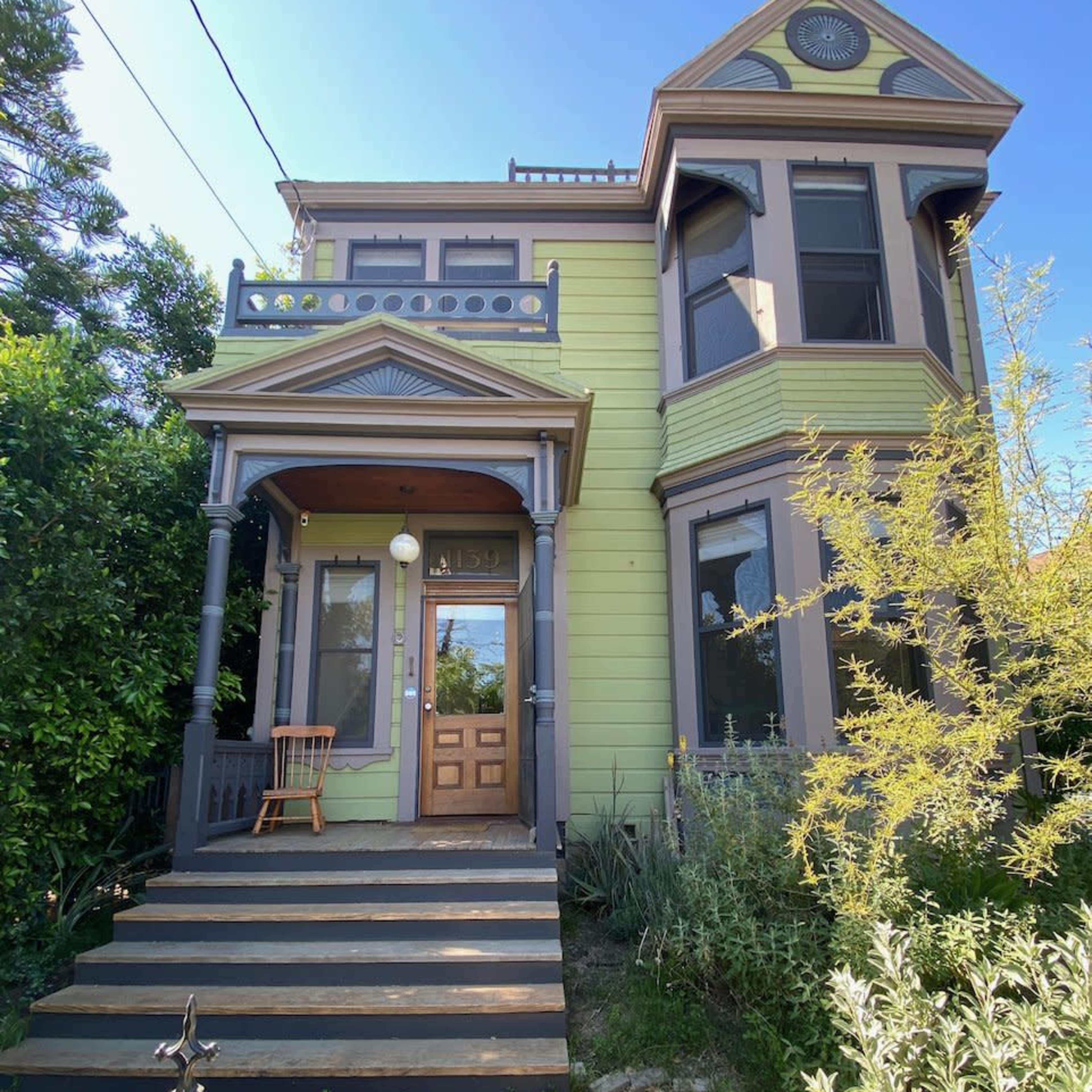 A two-story Victorian-style house with light green siding, a brown front door, and a porch supported by columns, surrounded by shrubs and a picket fence.
