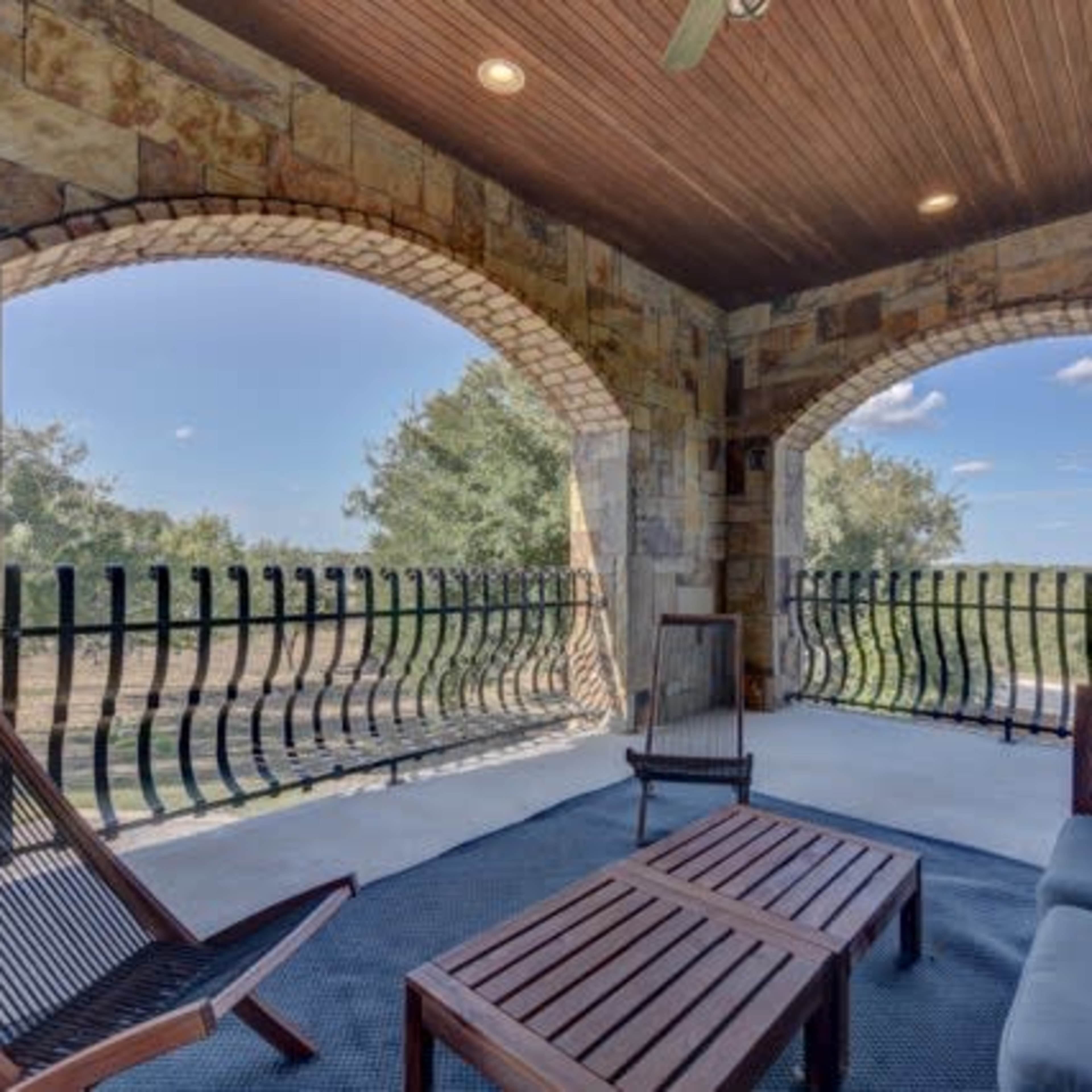 A covered patio with stone walls and arched openings overlooking a grassy landscape, furnished with wooden seating and a small table.