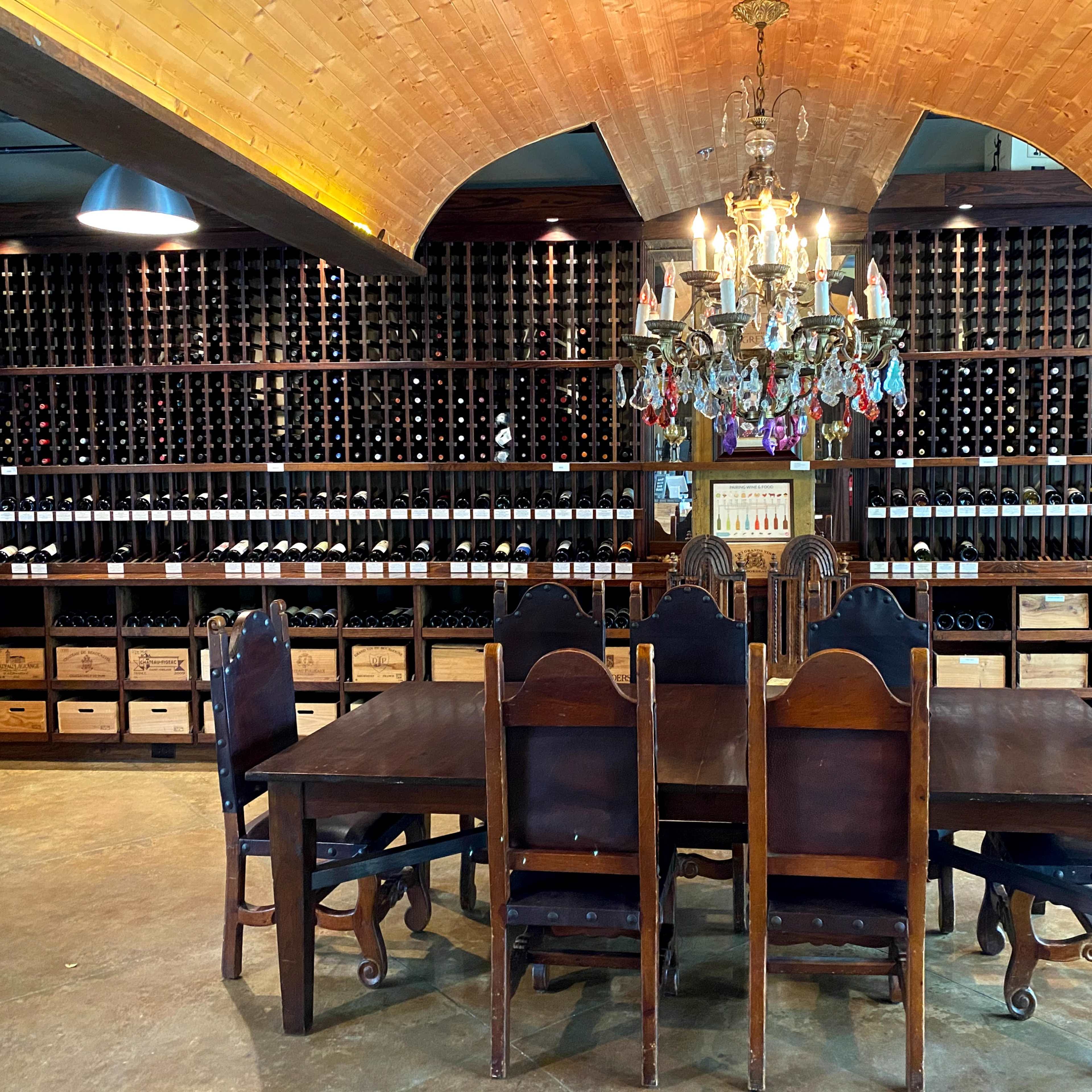 A large wooden dining table with sturdy chairs is set in front of a wall filled with neatly arranged wine bottles and a chandelier overhead.