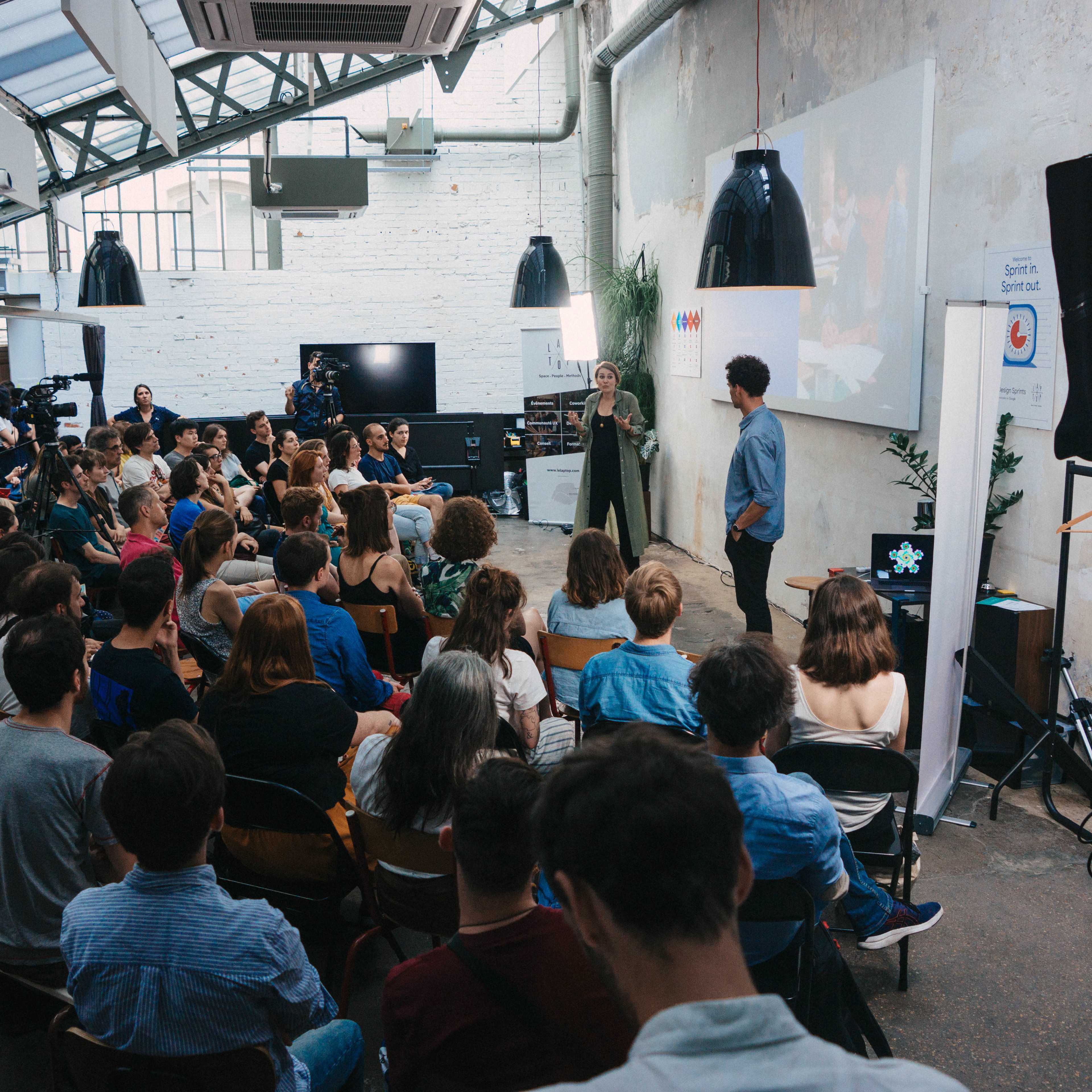 A crowd of people listens attentively to two presenters on stage at a well-lit event in an industrial-style venue.