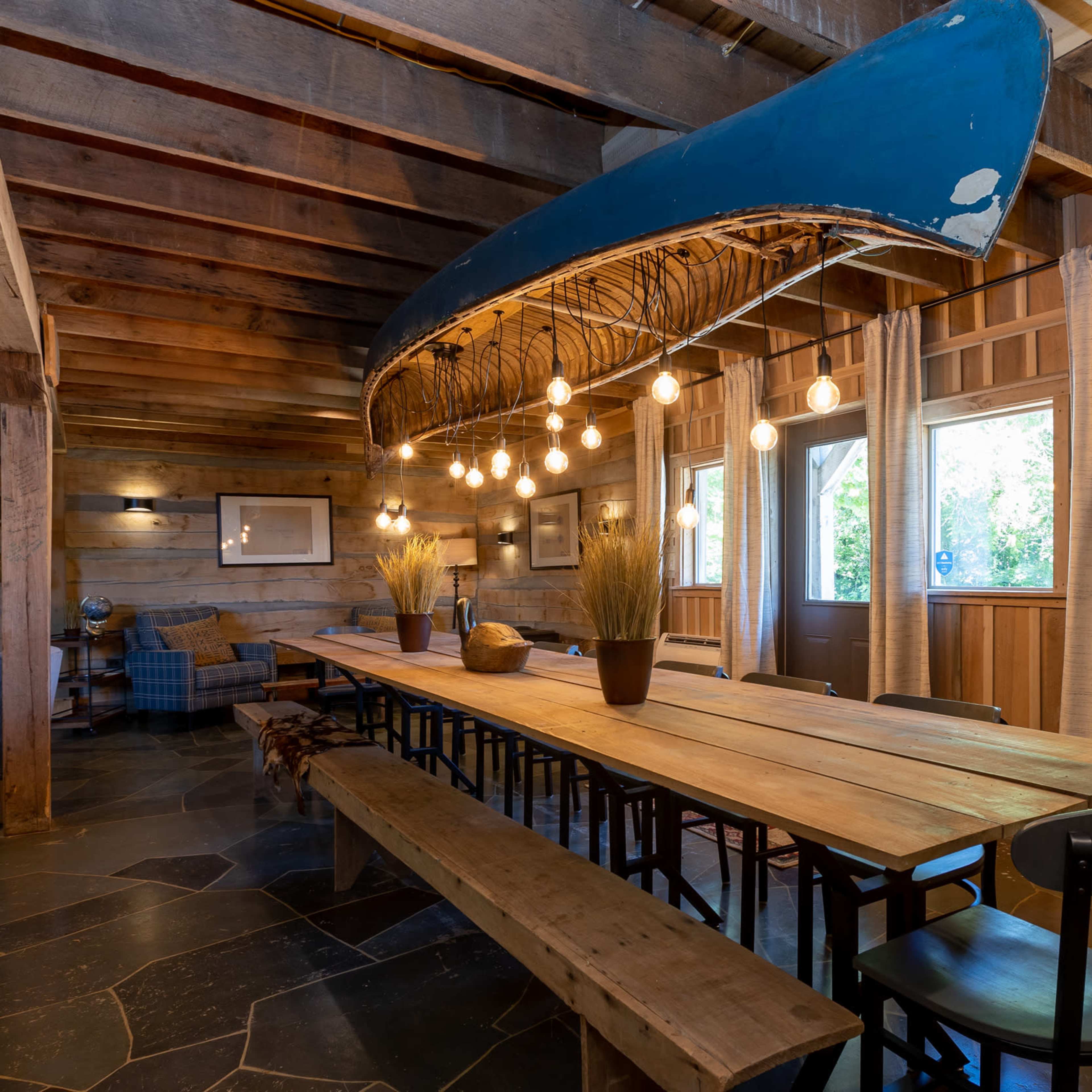 A rustic dining area features a long wooden table with benches, illuminated by pendant lights suspended from a canoe above.