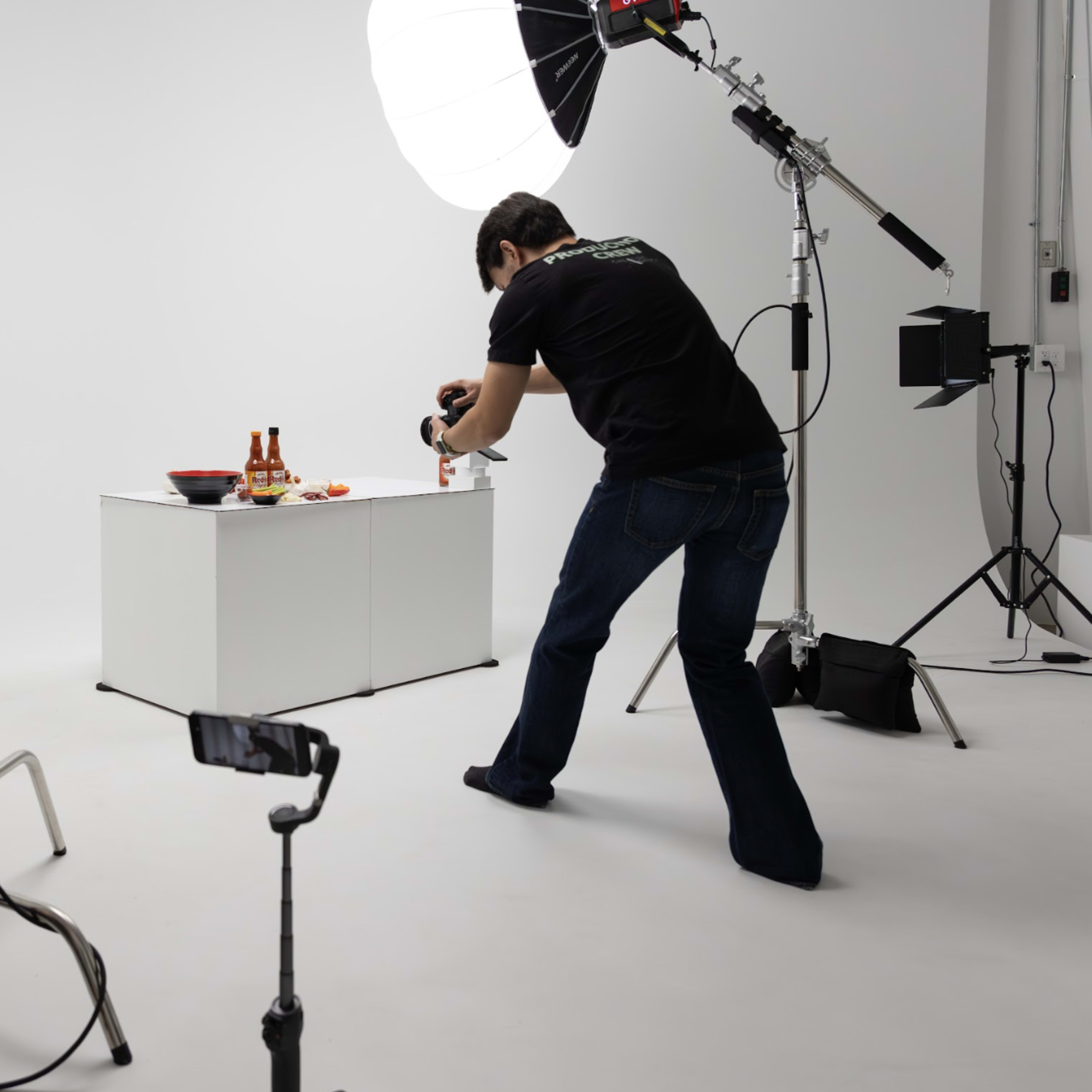 A photographer wearing a black shirt captures an image of food on a table in a studio setting with multiple lighting sources.