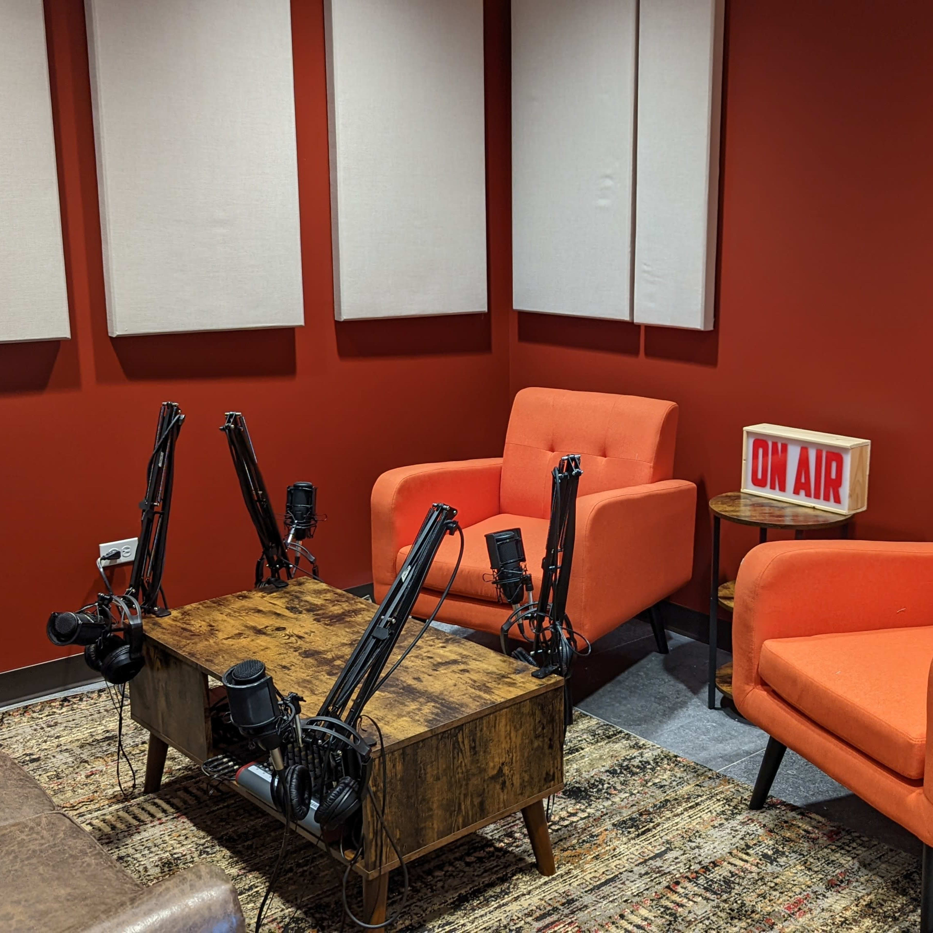 A recording studio with two orange chairs, a wooden table, and a lit "ON AIR" sign against a red wall.