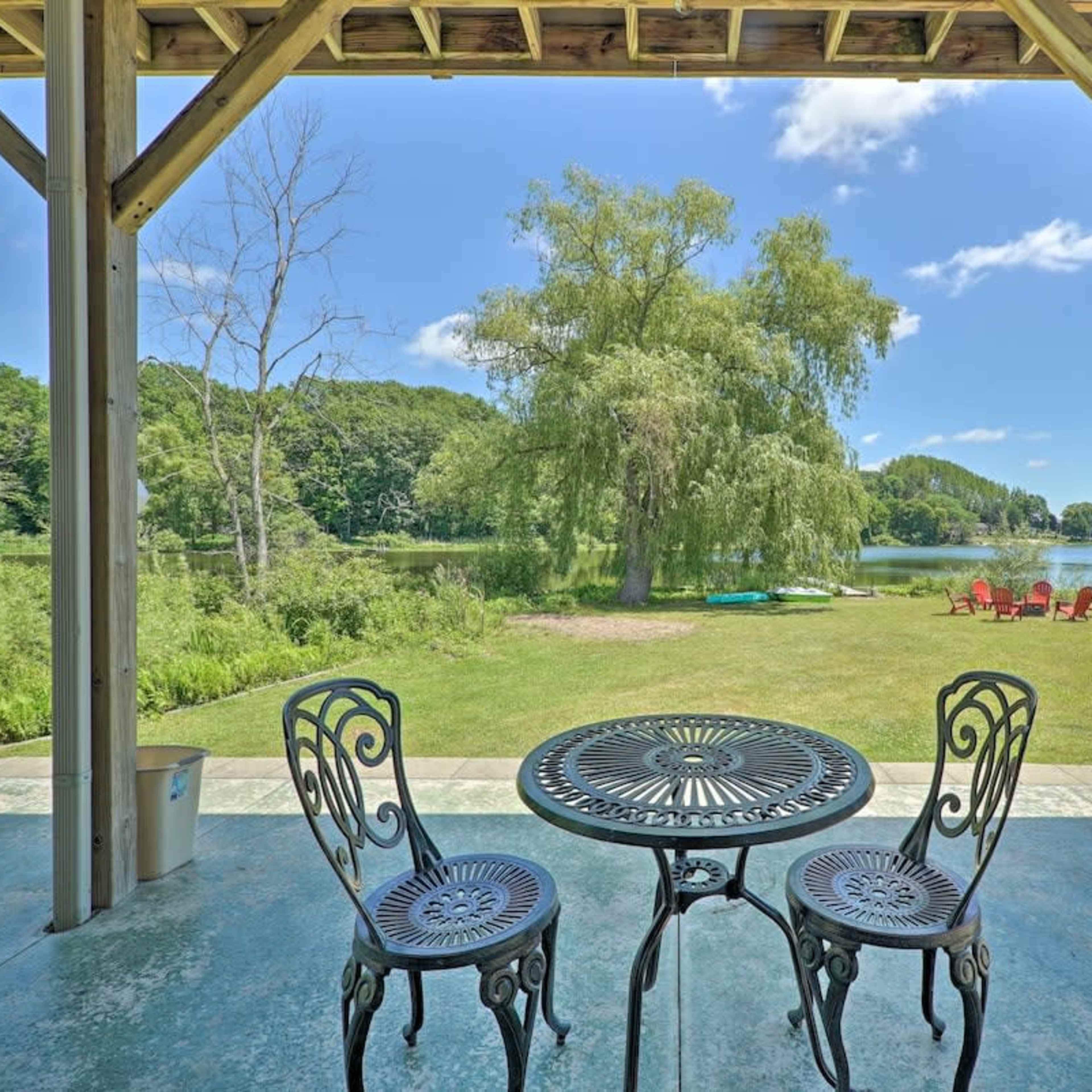 A metal table and two chairs are placed under a covered porch, overlooking a grassy area and a lake with trees in the background.