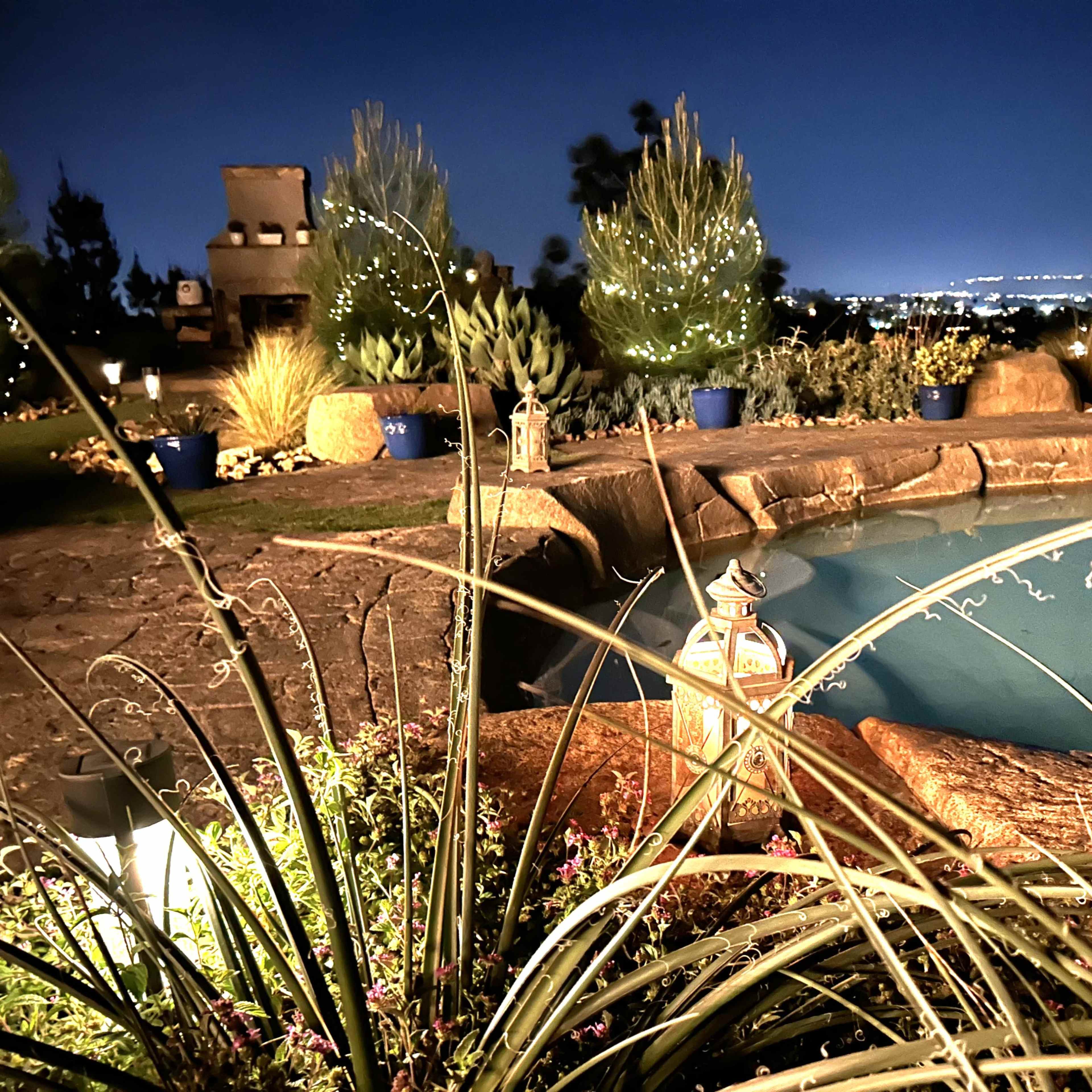 A illuminated backyard pool area at night, surrounded by greenery and decorative lanterns.