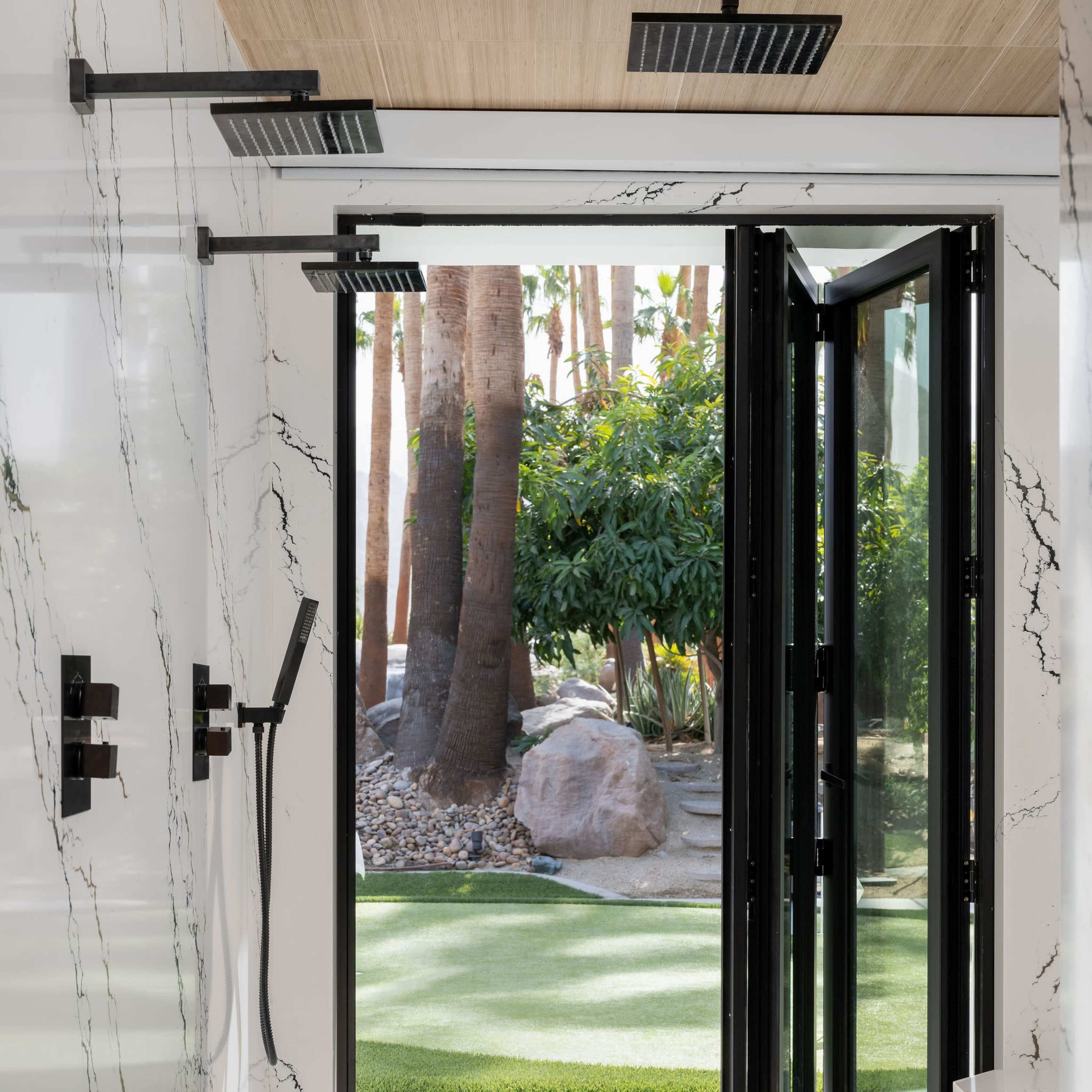 A modern bathroom features a glass door opening to a landscaped outdoor area with palm trees and a grass lawn.
