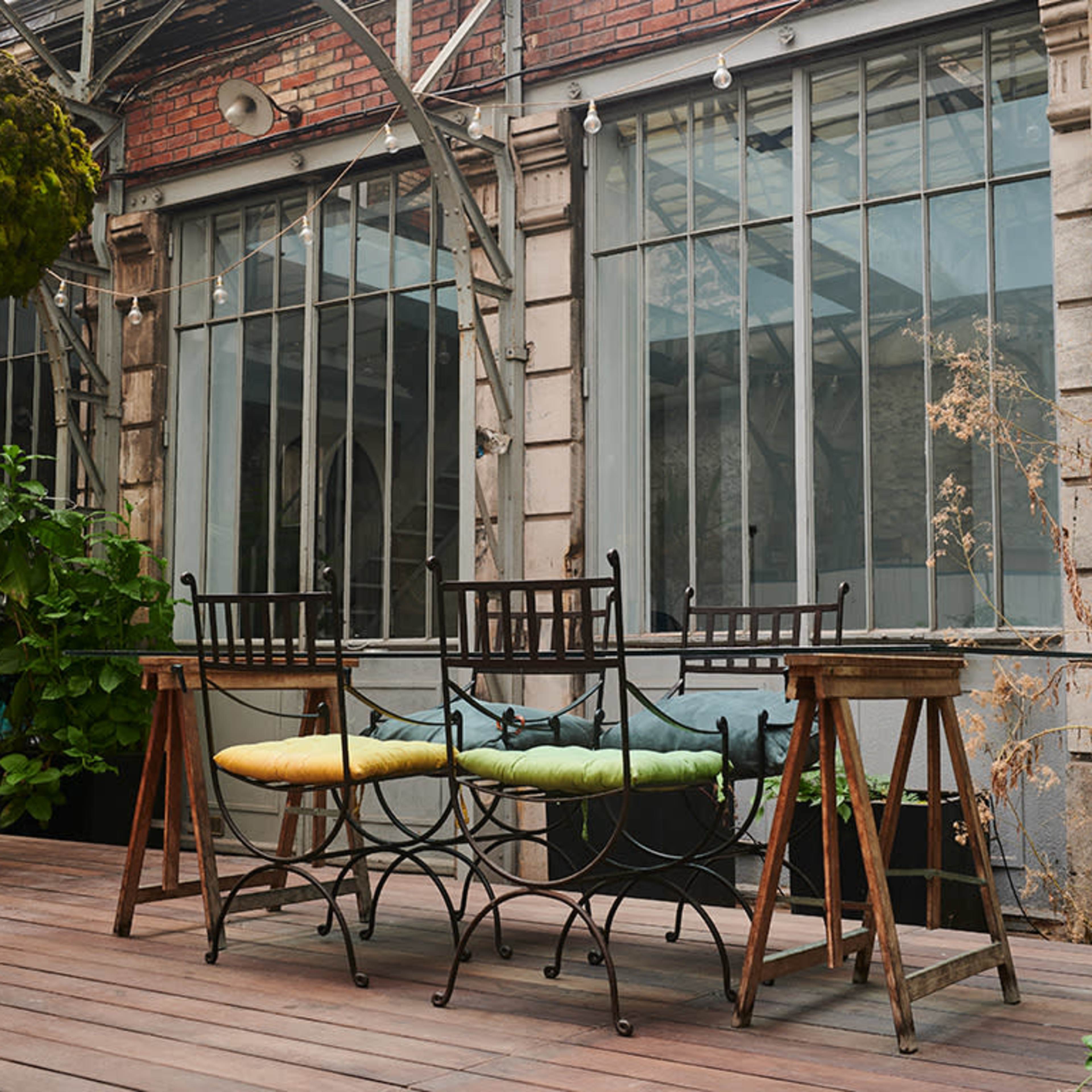 A wooden deck features a metal table with four chairs, surrounded by plants and large windows in a rustic building.
