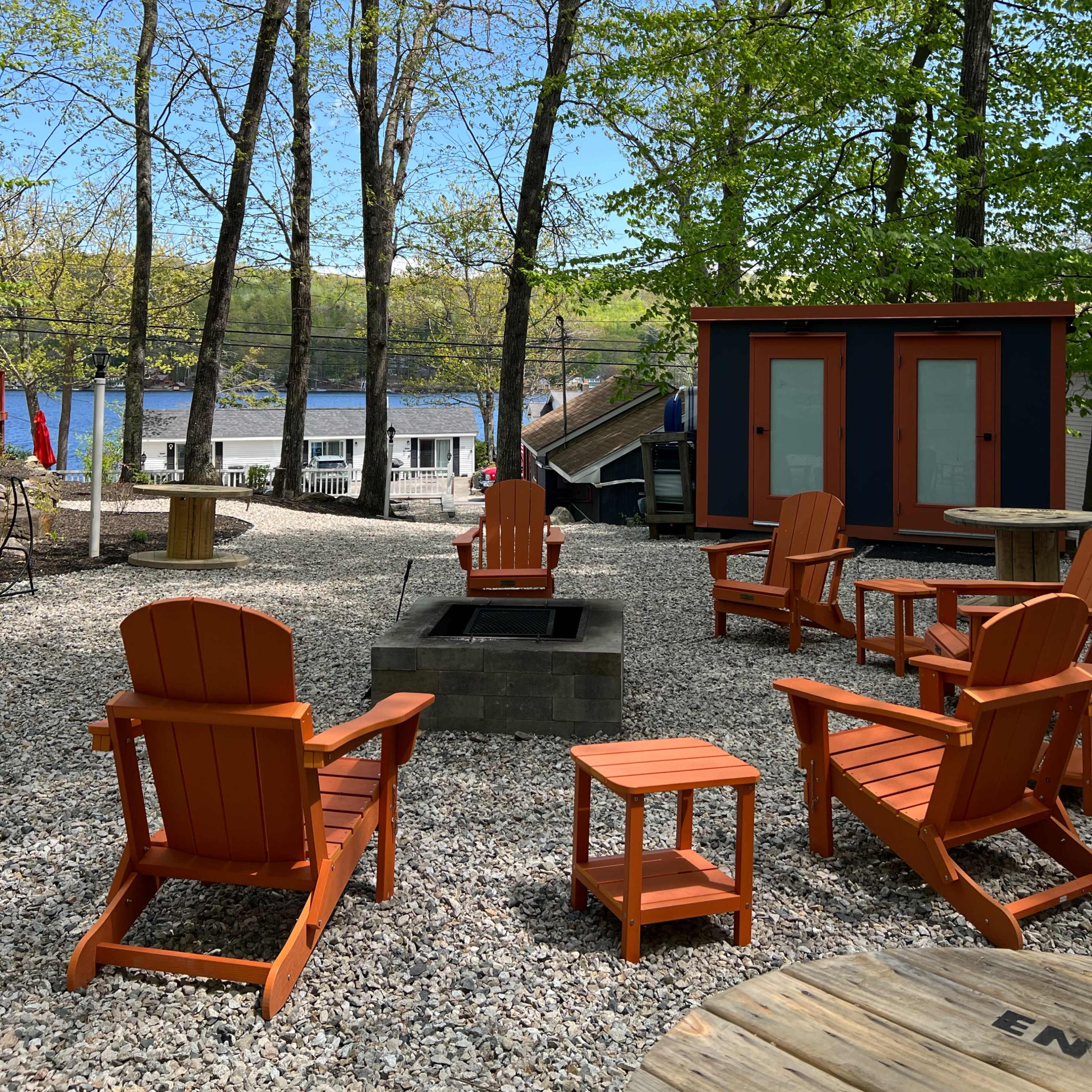 A seating area with orange Adirondack chairs and a small table is set among trees near a lake, featuring a fire pit and a small building in the background.
