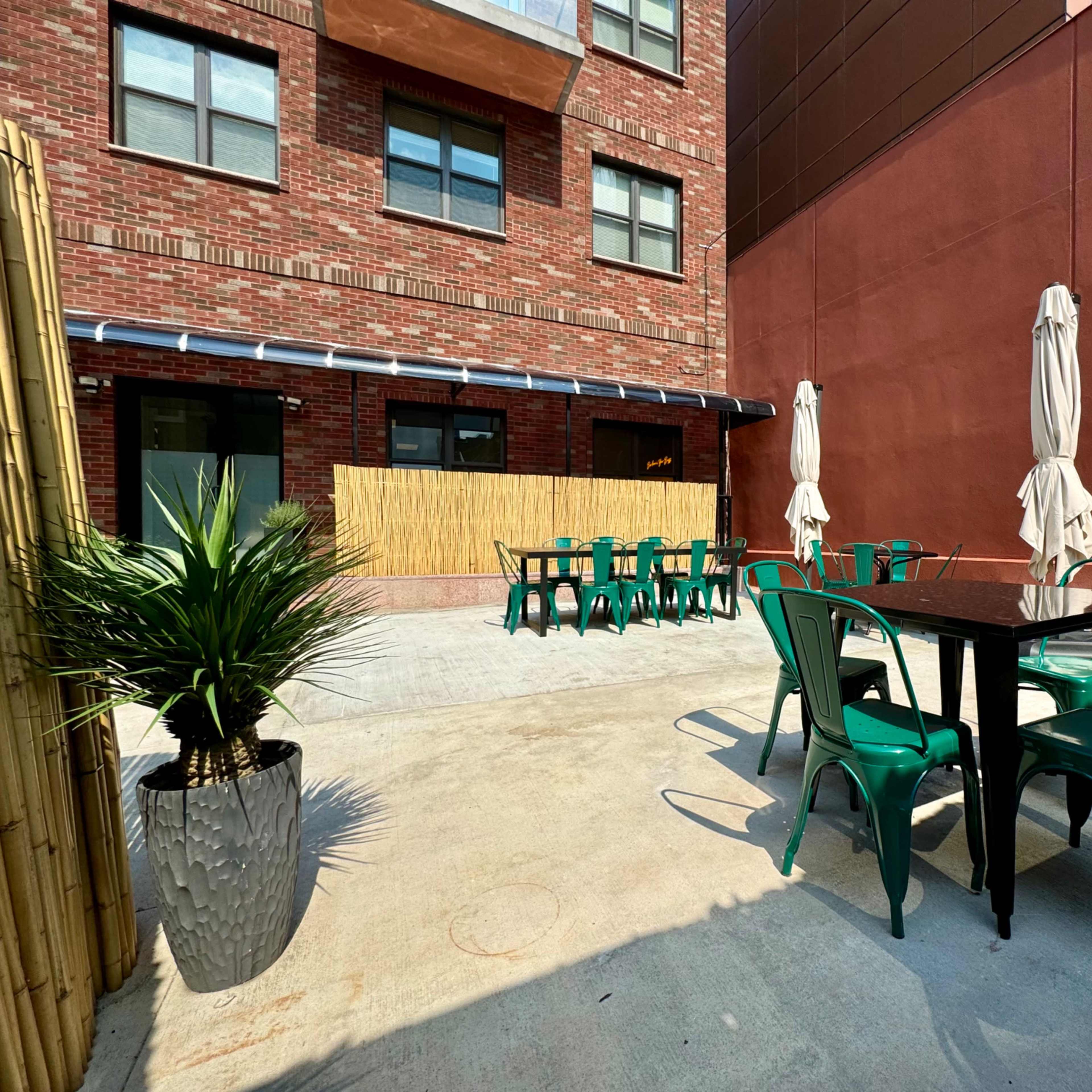 An outdoor seating area with green metal chairs and tables, surrounded by a brick building and bamboo screens.