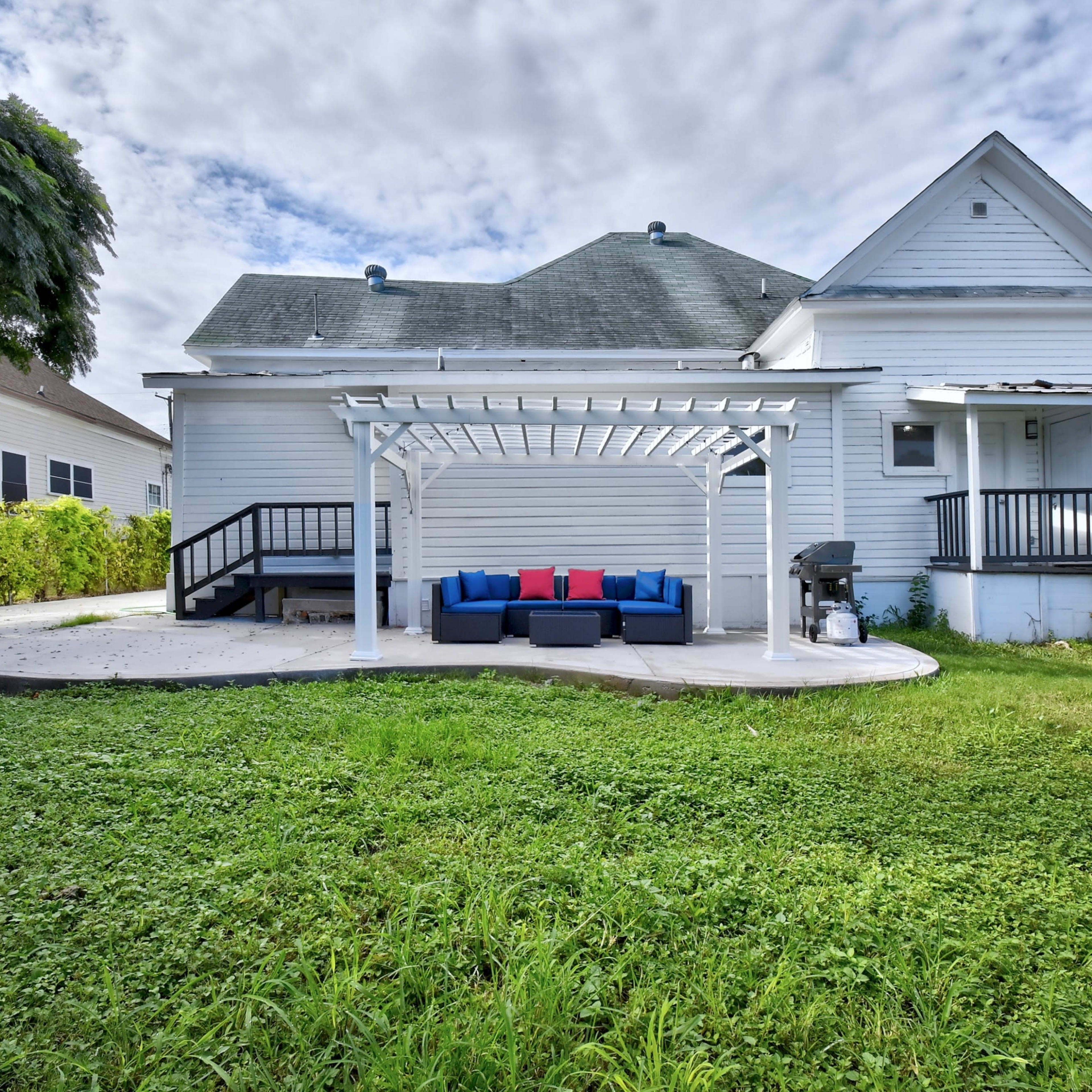The back of a white house with a covered patio featuring a blue and red outdoor sofa and a grill, surrounded by grass and small trees.