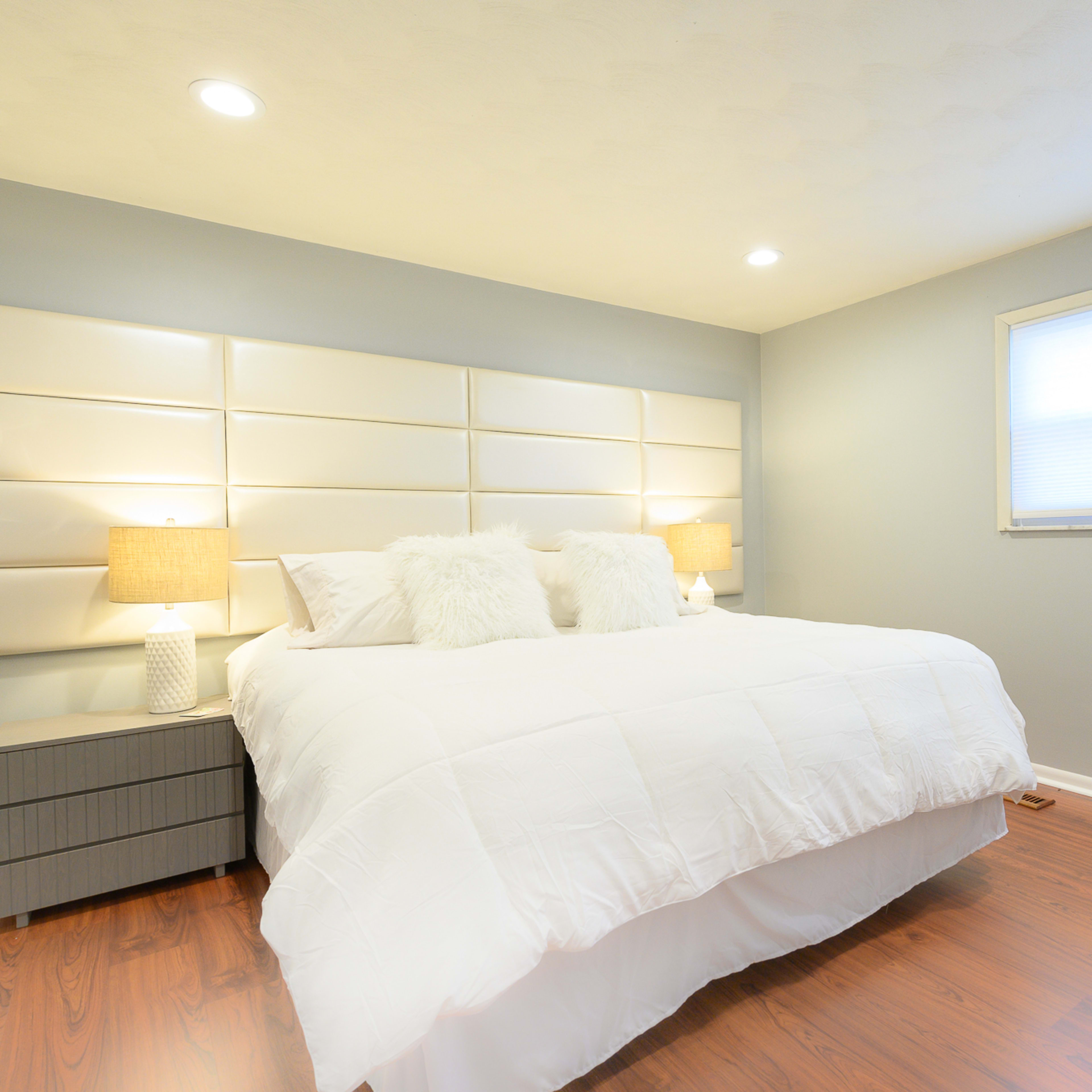 A well-lit bedroom features a large bed with white linens, two bedside tables with lamps, and a large window.