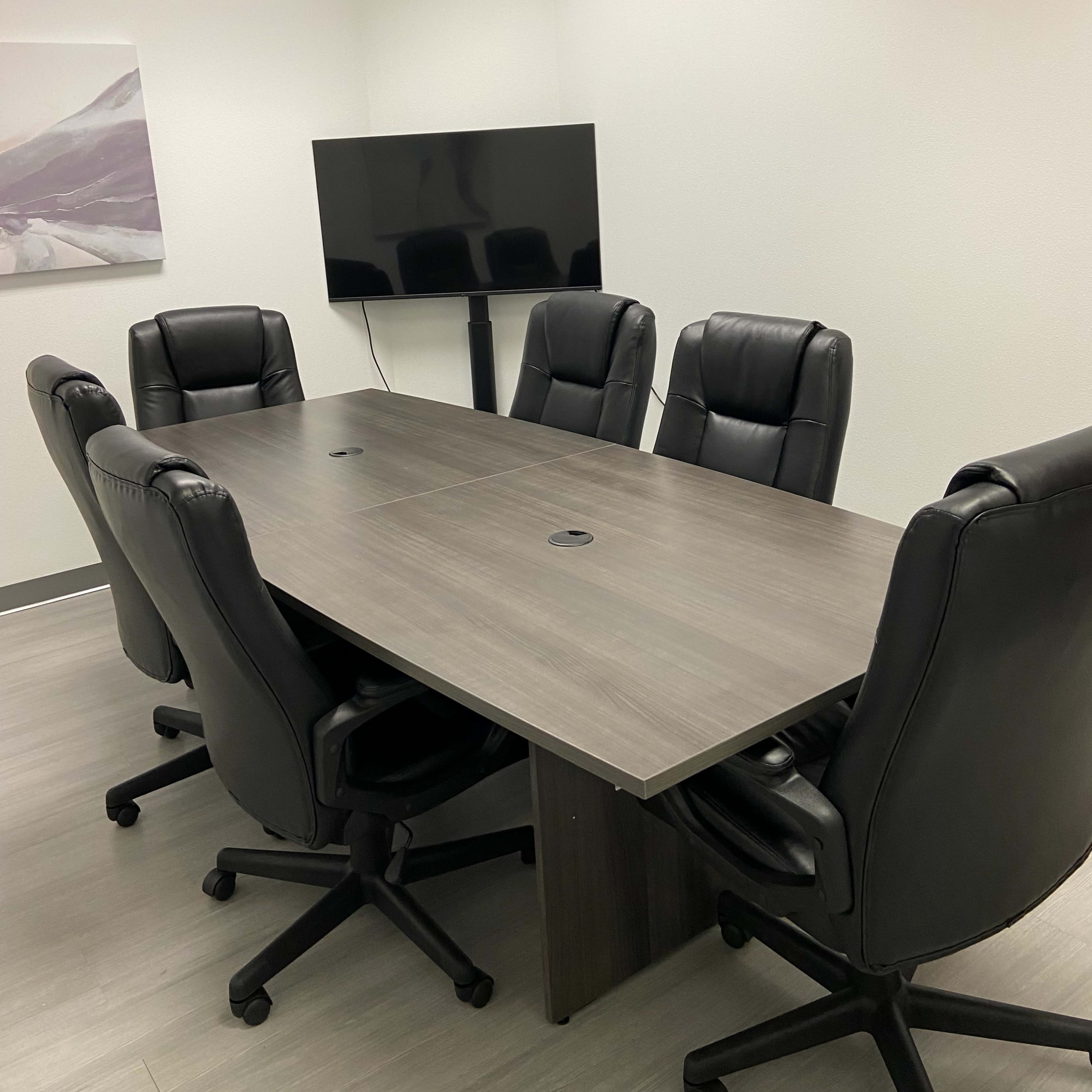A conference room features a large rectangular table surrounded by six black ergonomic chairs and a wall-mounted television.
