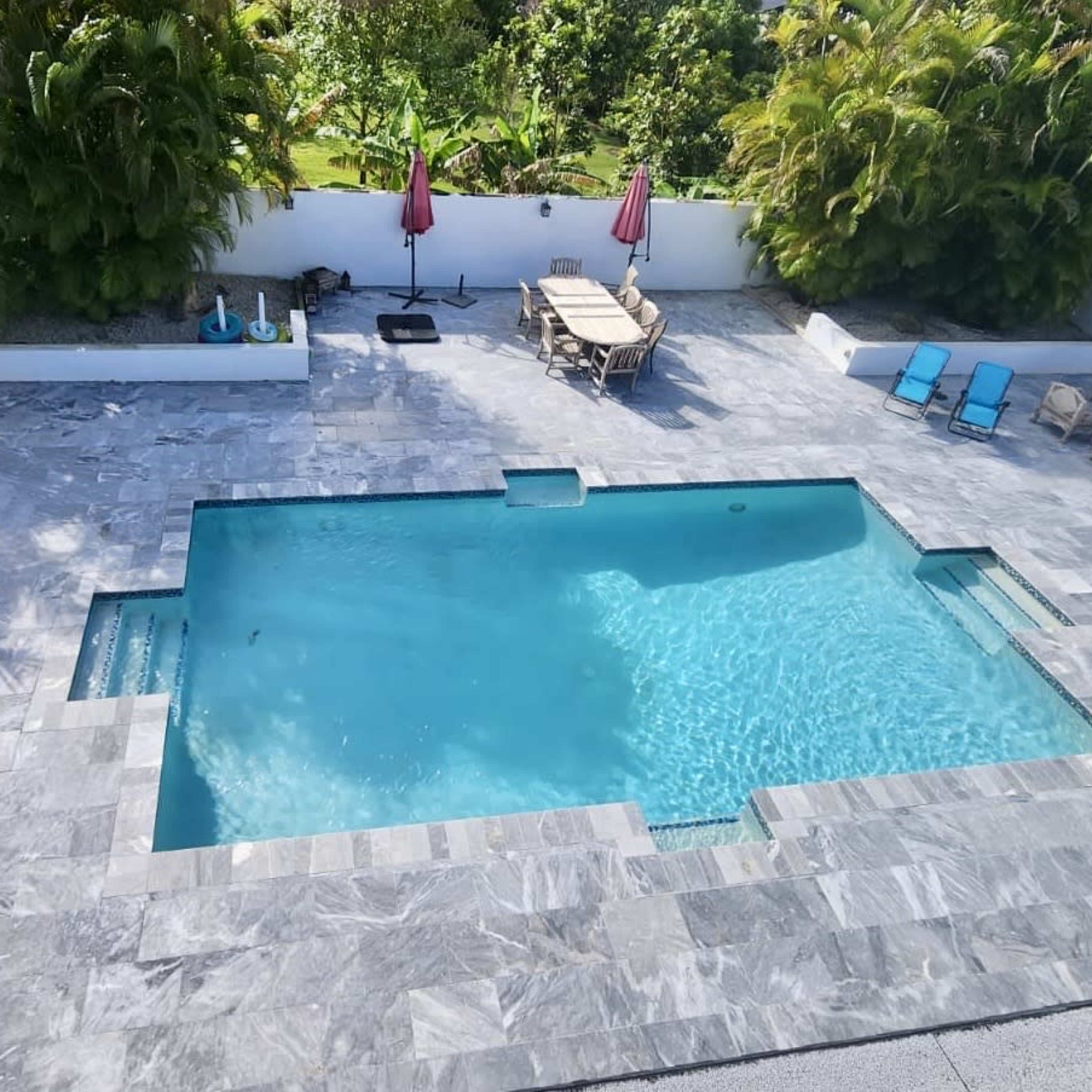 A rectangular swimming pool surrounded by a stone patio, with lounge chairs and umbrellas nearby.