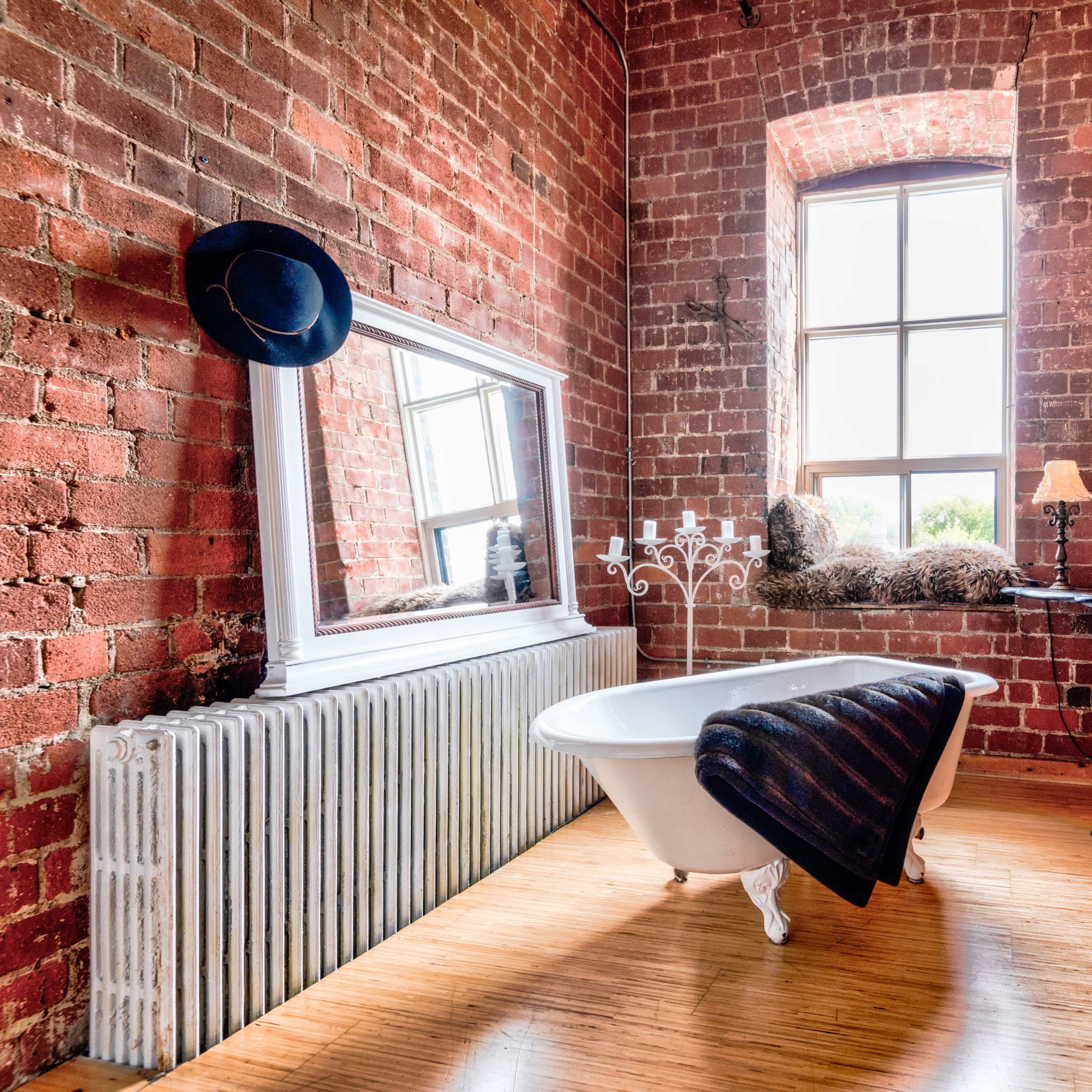 A rustic room featuring a freestanding bathtub, a large mirror, a table with a lamp, and a radiator against exposed brick walls.