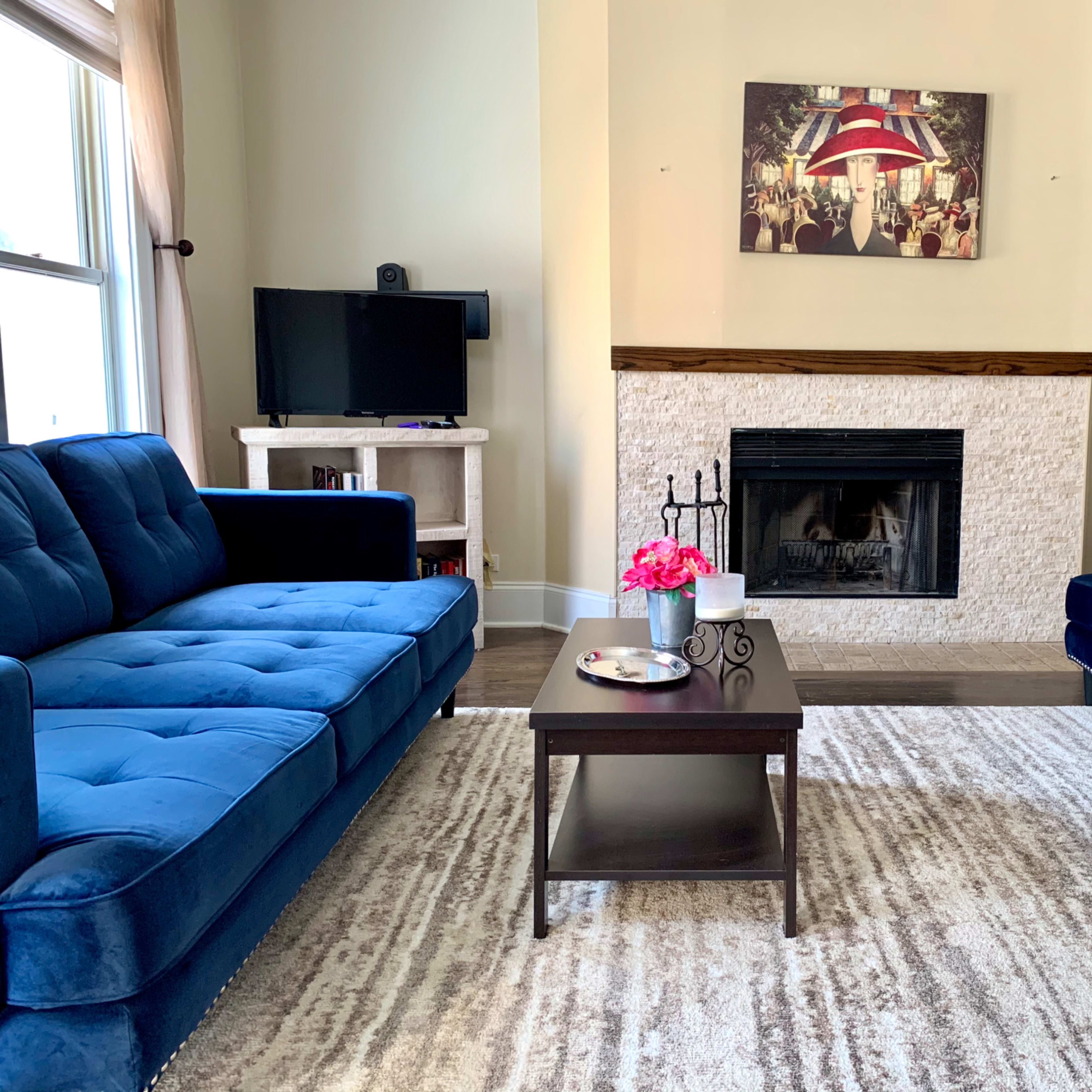 The living room features two blue sofas facing a coffee table, a fireplace, and a wall-mounted television under a framed artwork.