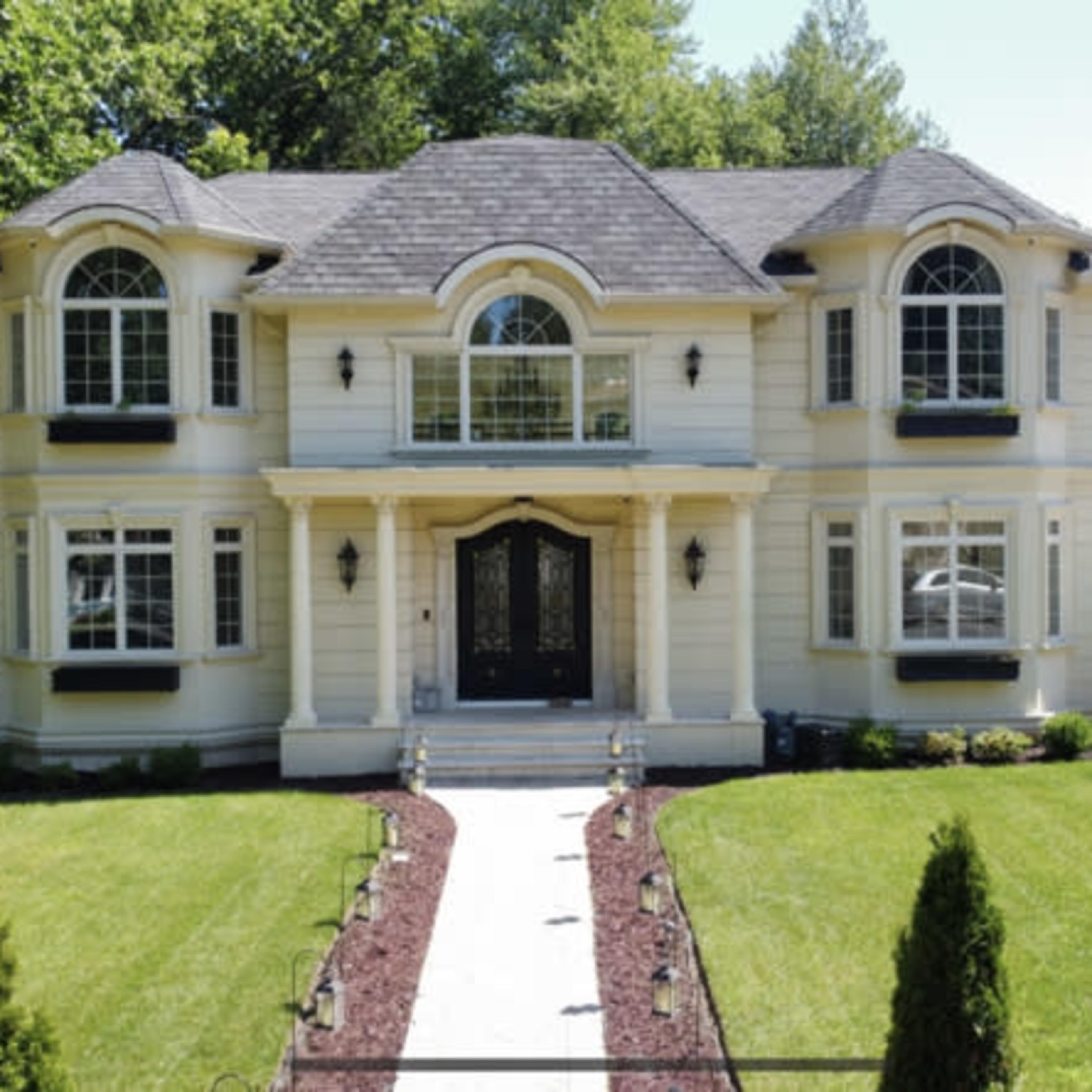 A two-story house with white siding, large windows, and a well-manicured lawn, accompanied by a paved walkway leading to the entrance.