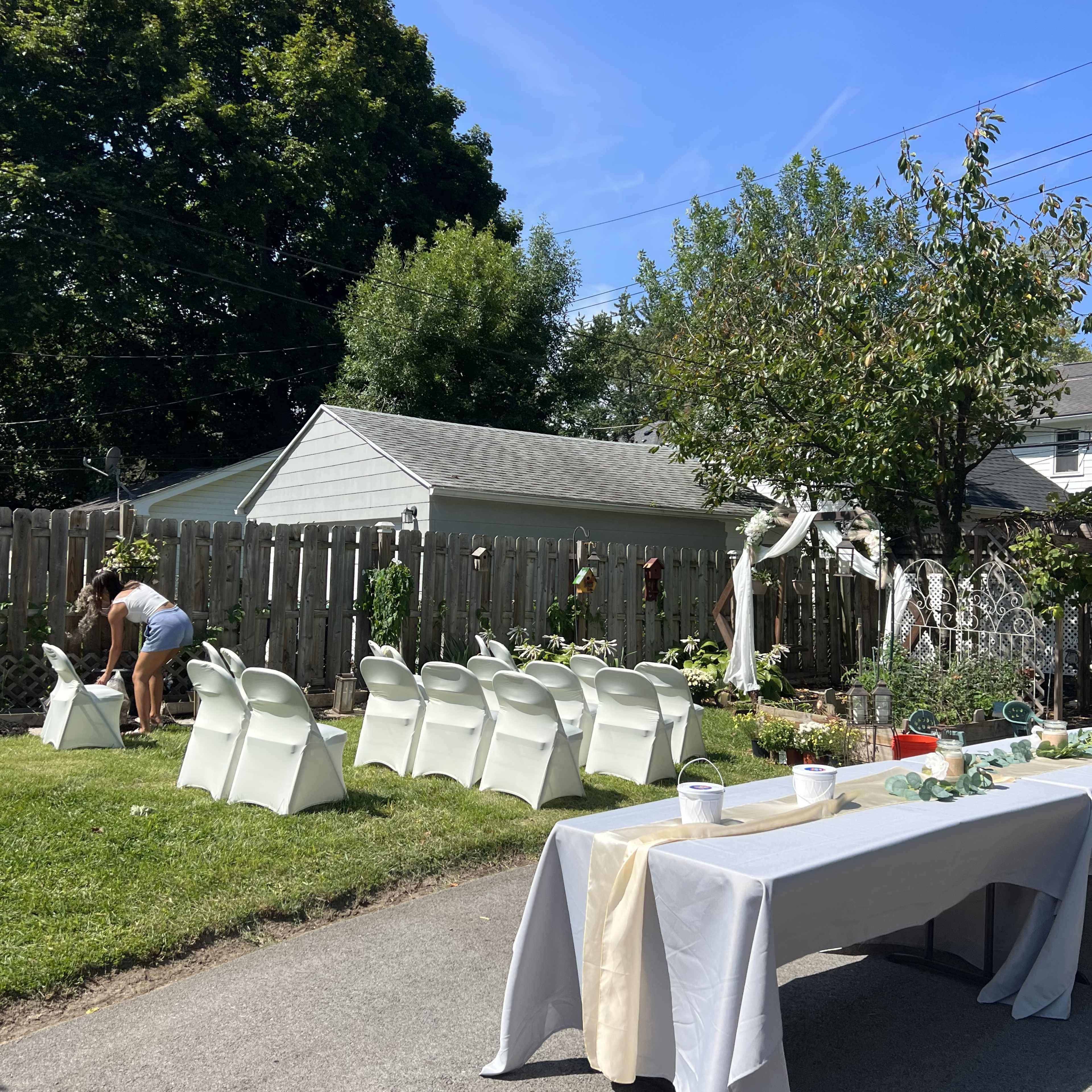 A backyard is set up for an event, featuring white chairs arranged in rows and a table adorned with a beige tablecloth, while a person adjusts decorations nearby.